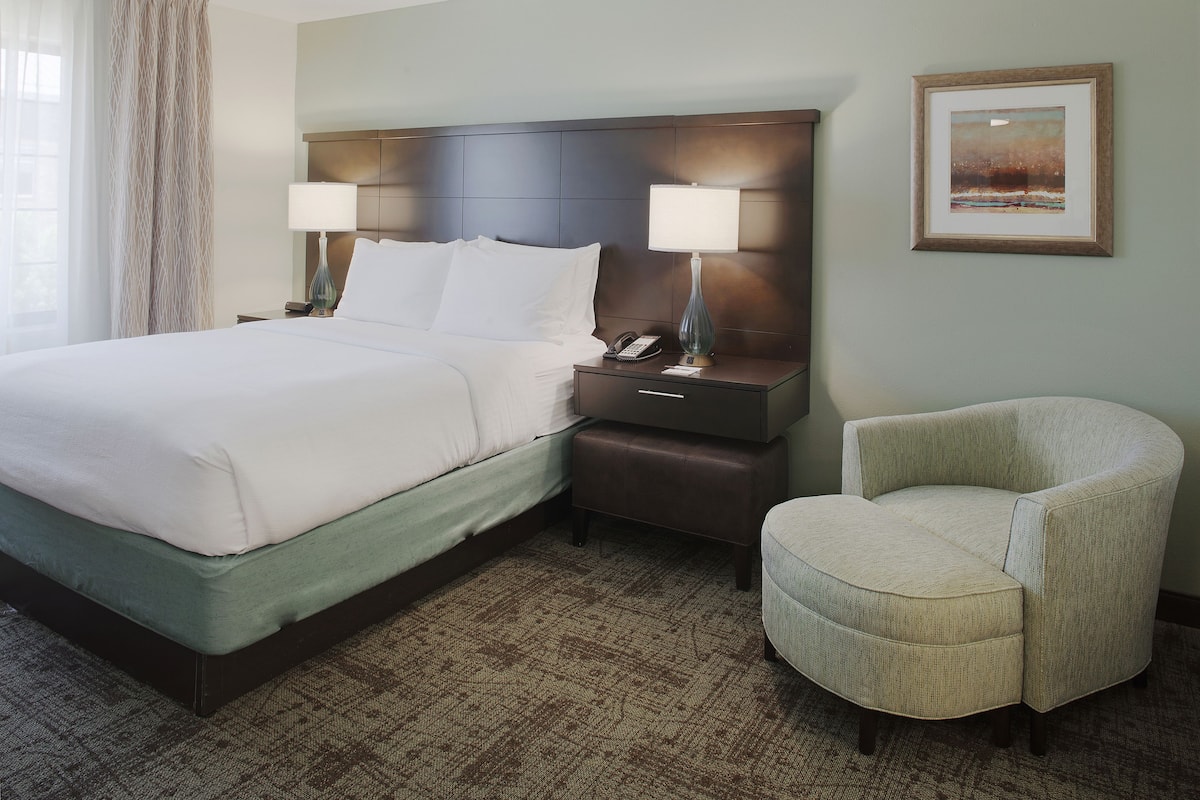 A hotel room with a bed, bedside lamps, a chair, a nightstand, and a framed picture on the wall.