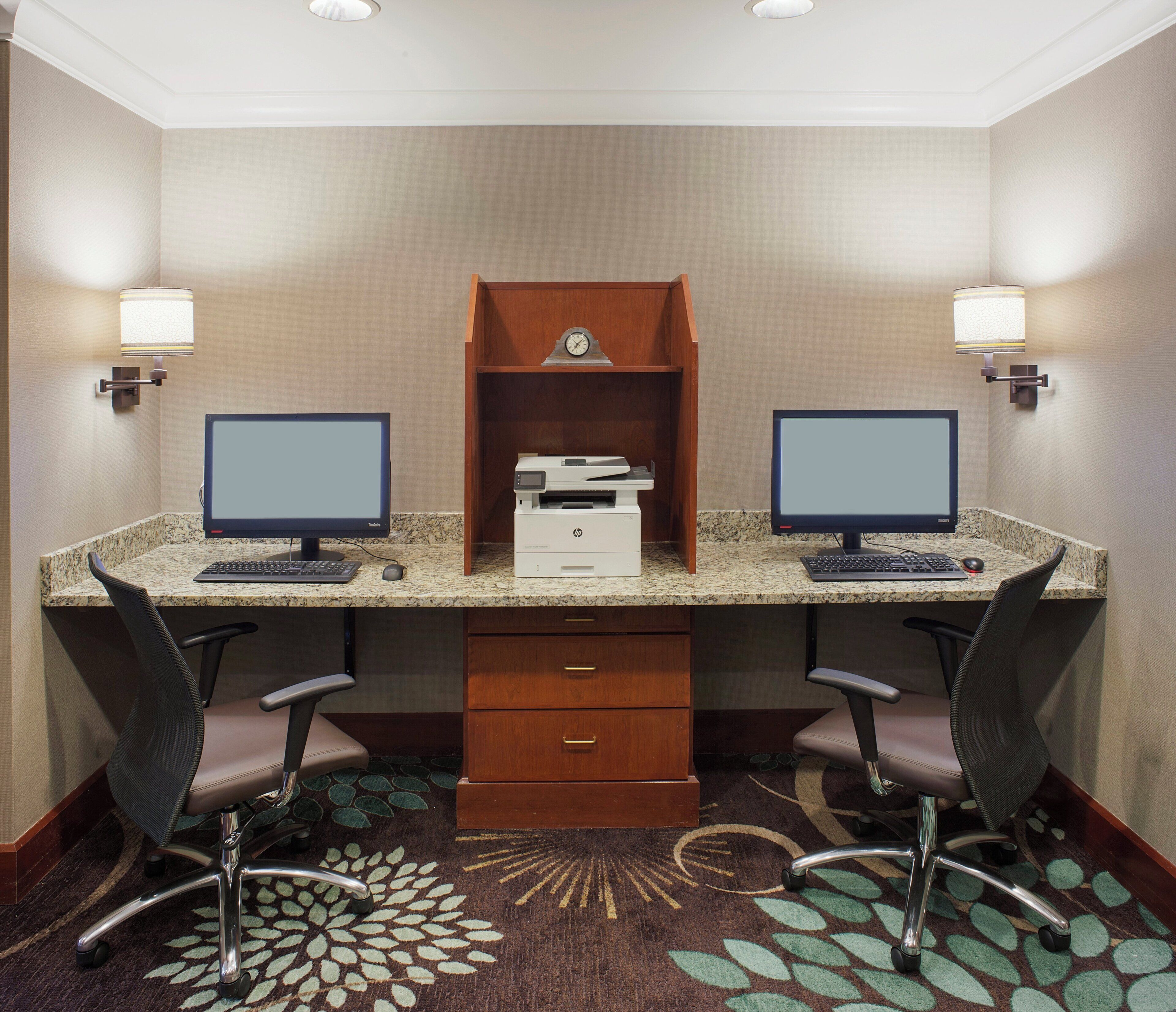 A hotel room with a desk, two monitors, a printer, and a clock on the wall.