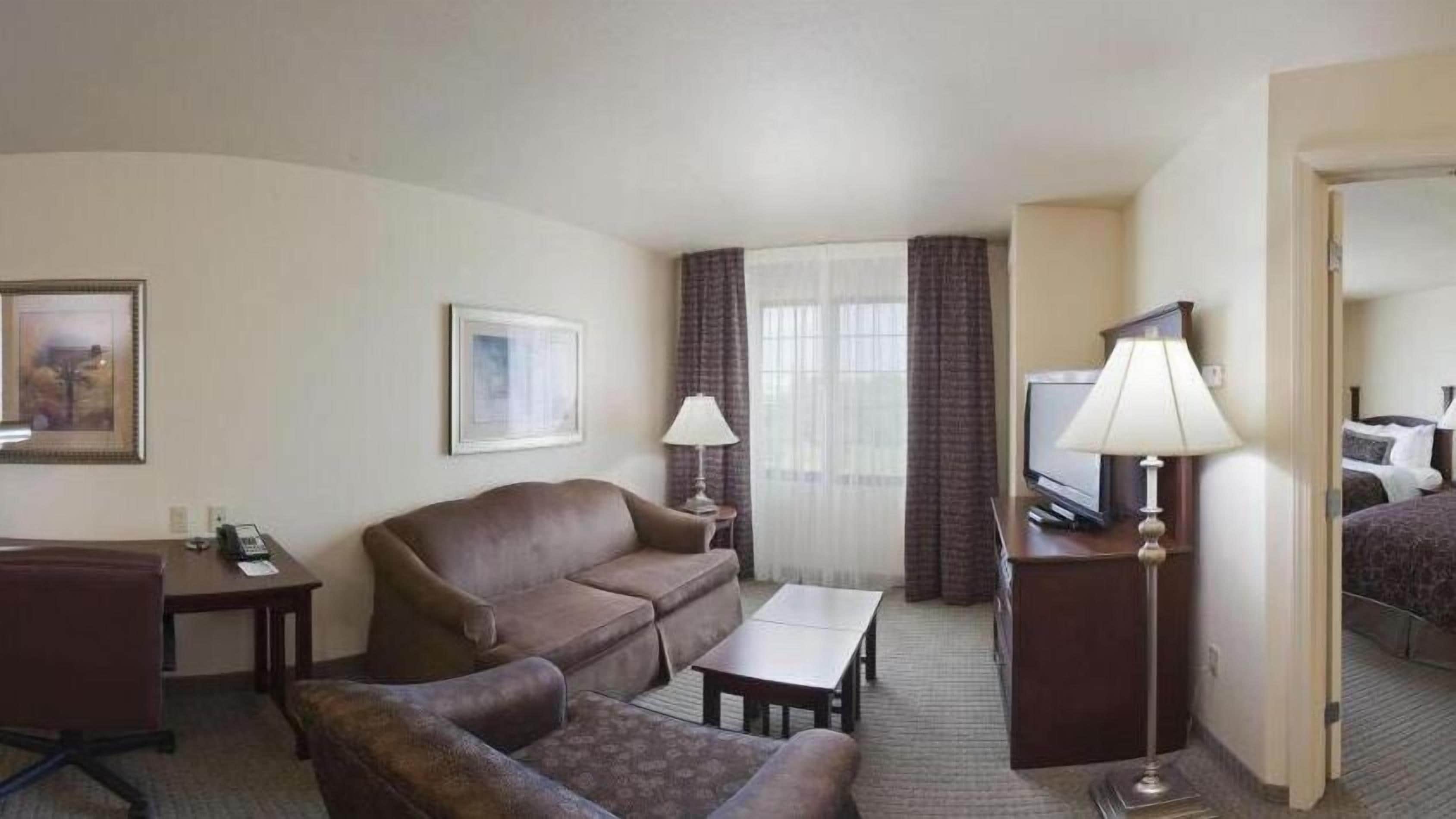 A hotel room with a brown sofa, a small table, a TV, and a lamp.