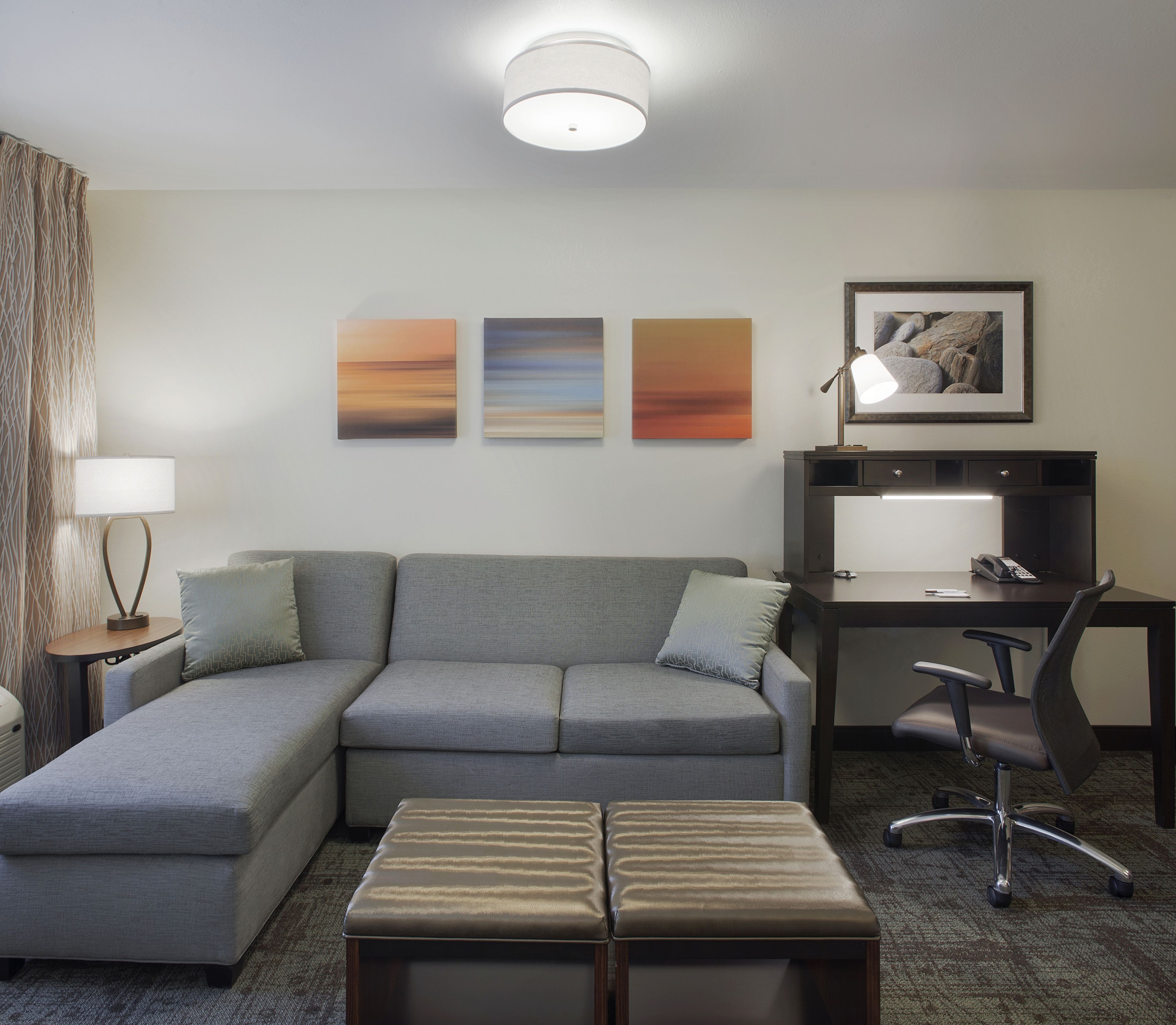 Habitación de hotel moderna con un sofá seccional gris, una mesita redonda con una lámpara, un escritorio con una silla y tres cuadros abstractos en la pared.
