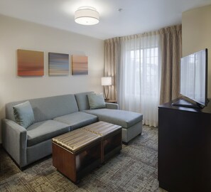 A modern living room with a grey sectional sofa, a wooden ottoman, a flat-screen TV, and three abstract wall art pieces. - Staybridge Suites Fayetteville/Univ Of Arkansas by IHG (Fayetteville)