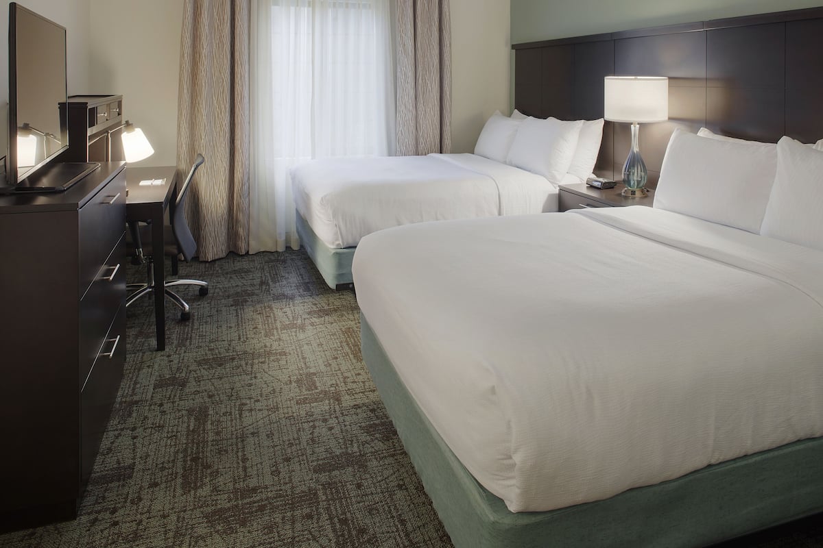 A hotel room with two beds, a desk, a chair, a lamp, and a large window with curtains.