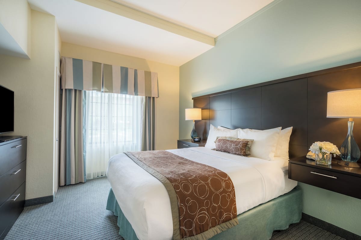 A hotel room with a large bed, a television, a desk with a lamp, and a window with curtains.