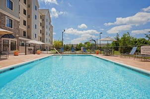 Piscina com água azul cristalina, rodeada por espreguiçadeiras e guarda-sóis, com um edifício ao fundo.