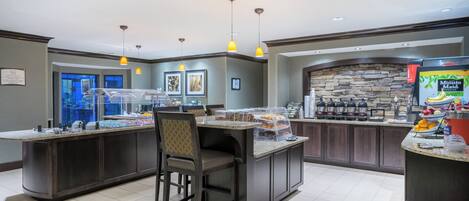 A hotel lobby with a self-service breakfast bar, seating area, and a stone accent wall.