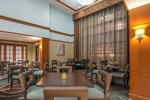 Uma área de refeições de hotel com várias mesas e cadeiras, uma janela grande com cortinas e um candeeiro fixado na parede.