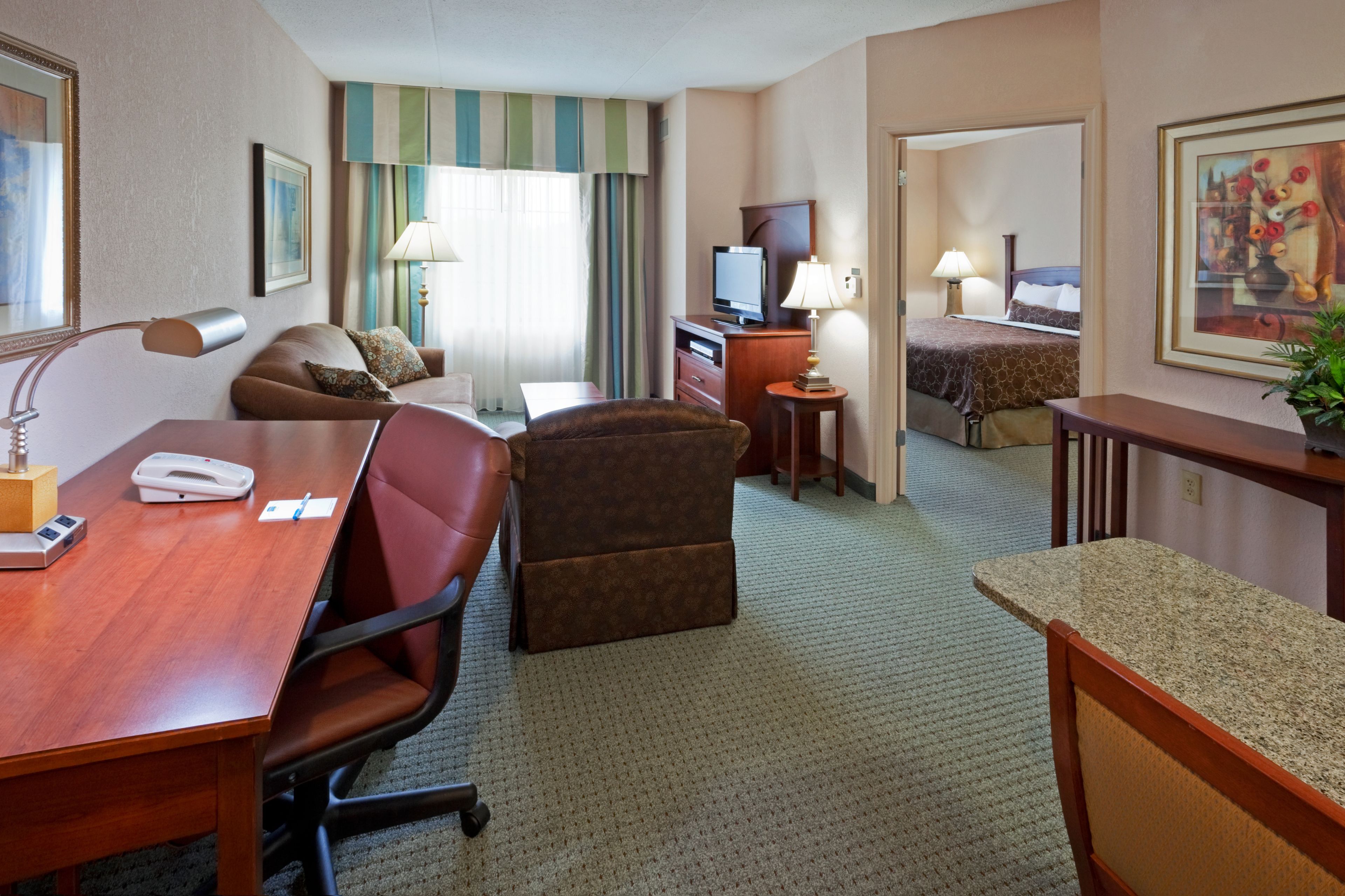A hotel room with a desk, a chair, a sofa, a bed, a television, and a lamp.