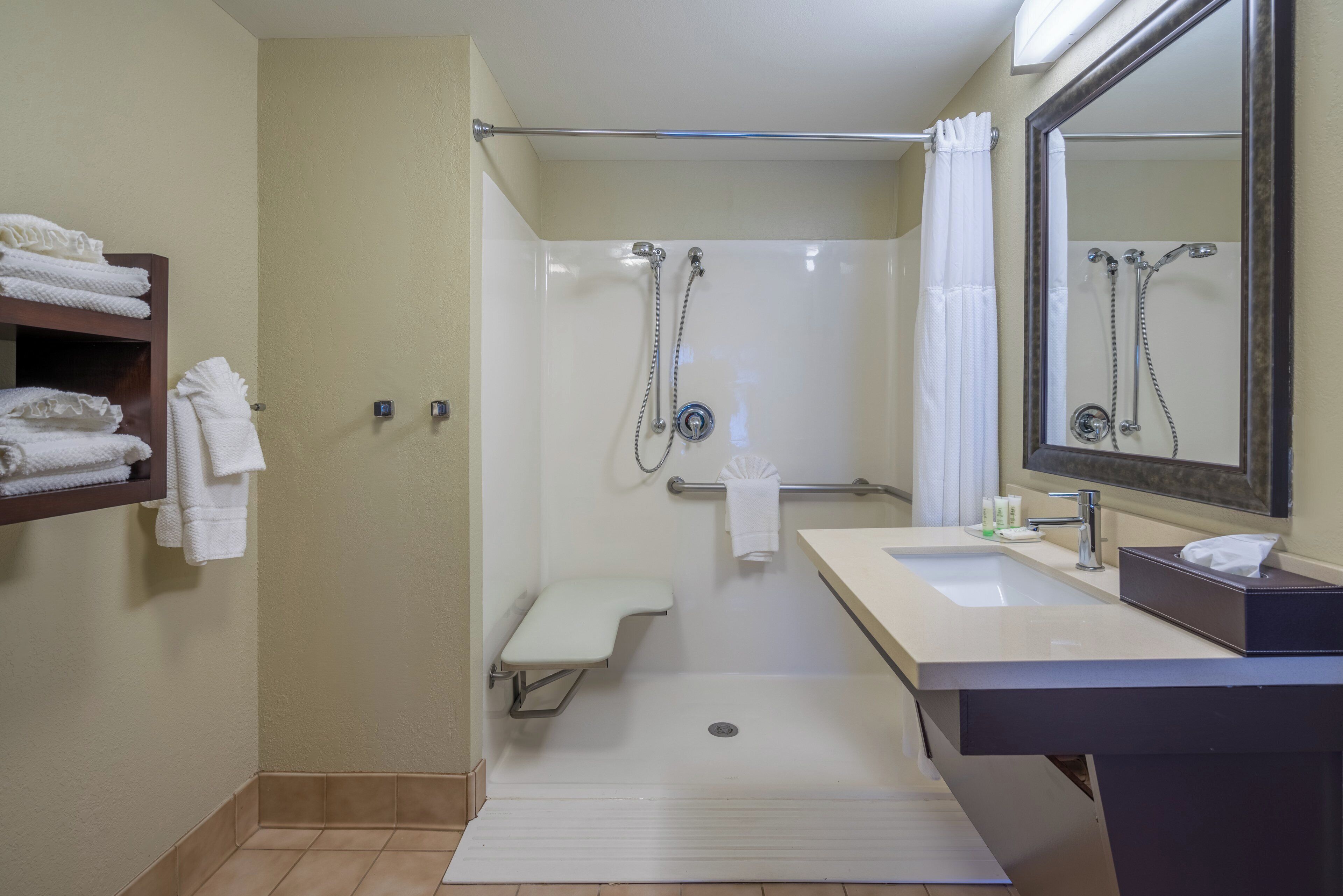 A standard hotel bathroom with a shower, a sink, a mirror, and a shelf with towels.