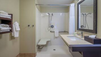 A standard hotel bathroom with a shower, a sink, a mirror, and a shelf with towels.
