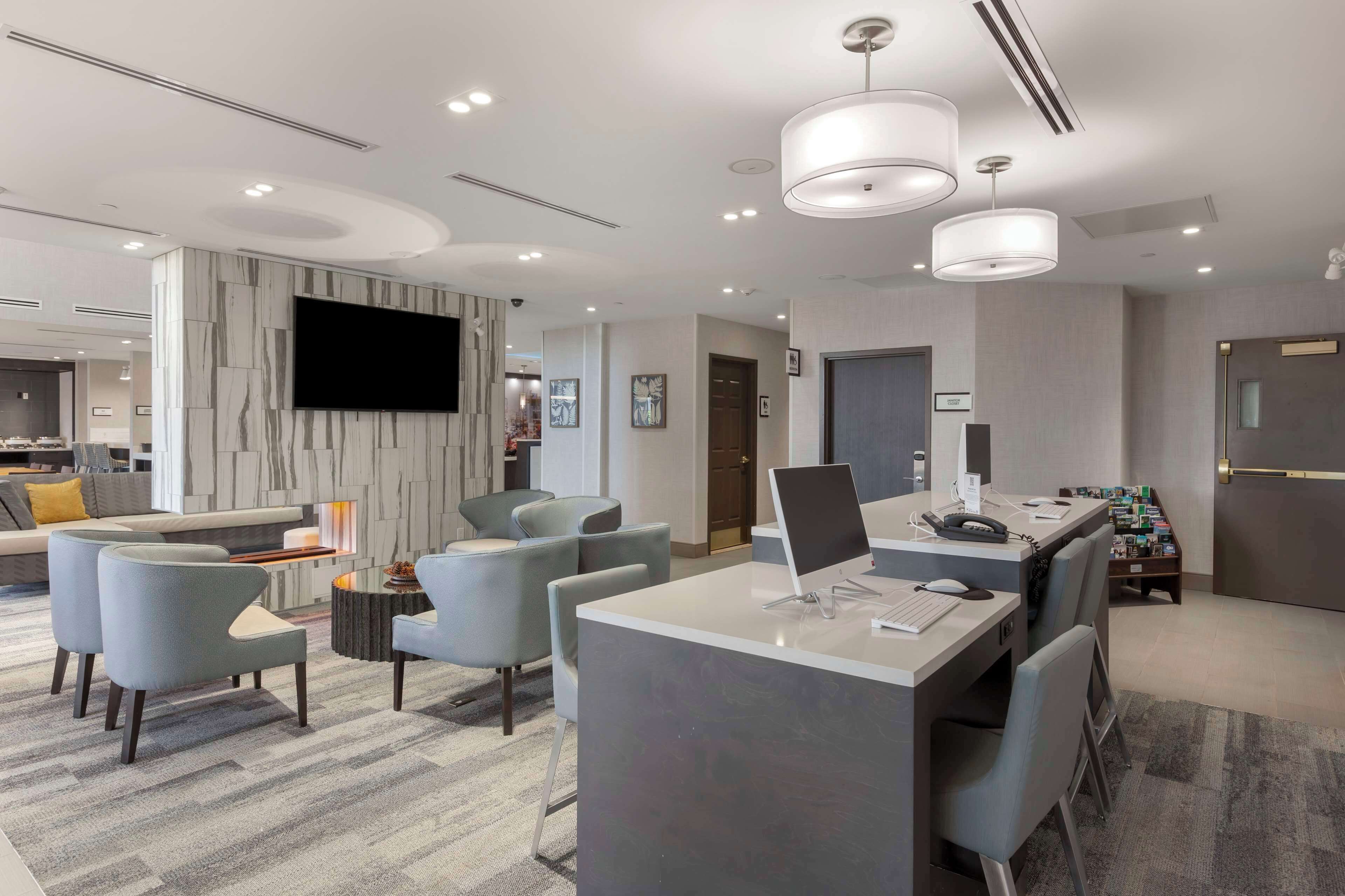 A modern hotel lobby with a seating area, a reception desk, and a large flat-screen TV mounted on the wall.