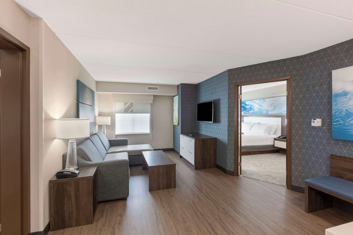 Una habitación de hotel moderna con sala de estar, dormitorio independiente, televisor de pantalla plana y un amplio ventanal.