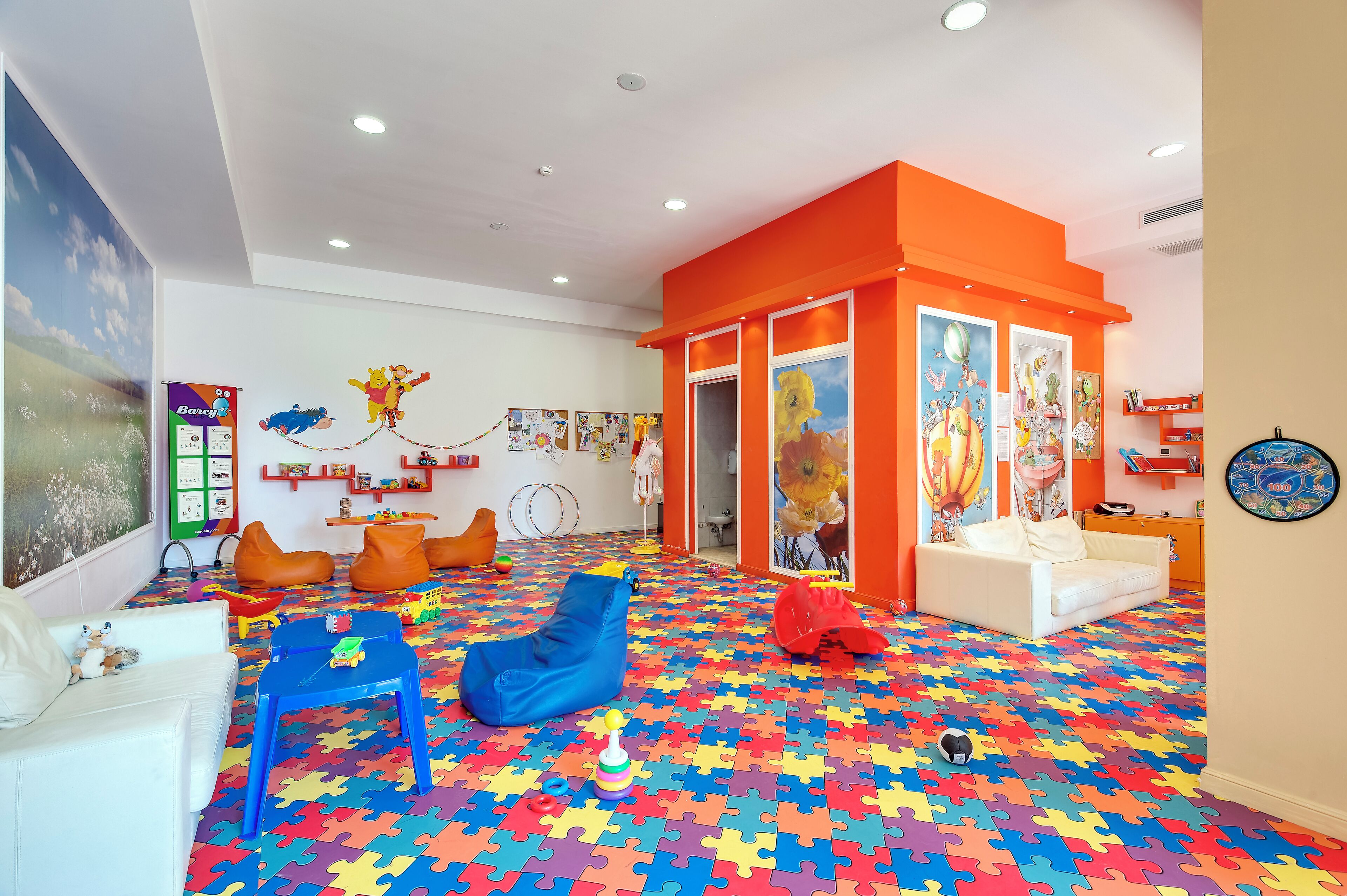 Un vibrante cuarto de juegos infantil con un piso de patrón de rompecabezas lleno de color, un sofá blanco, pufs naranjas y un sillón tipo bean bag azul.