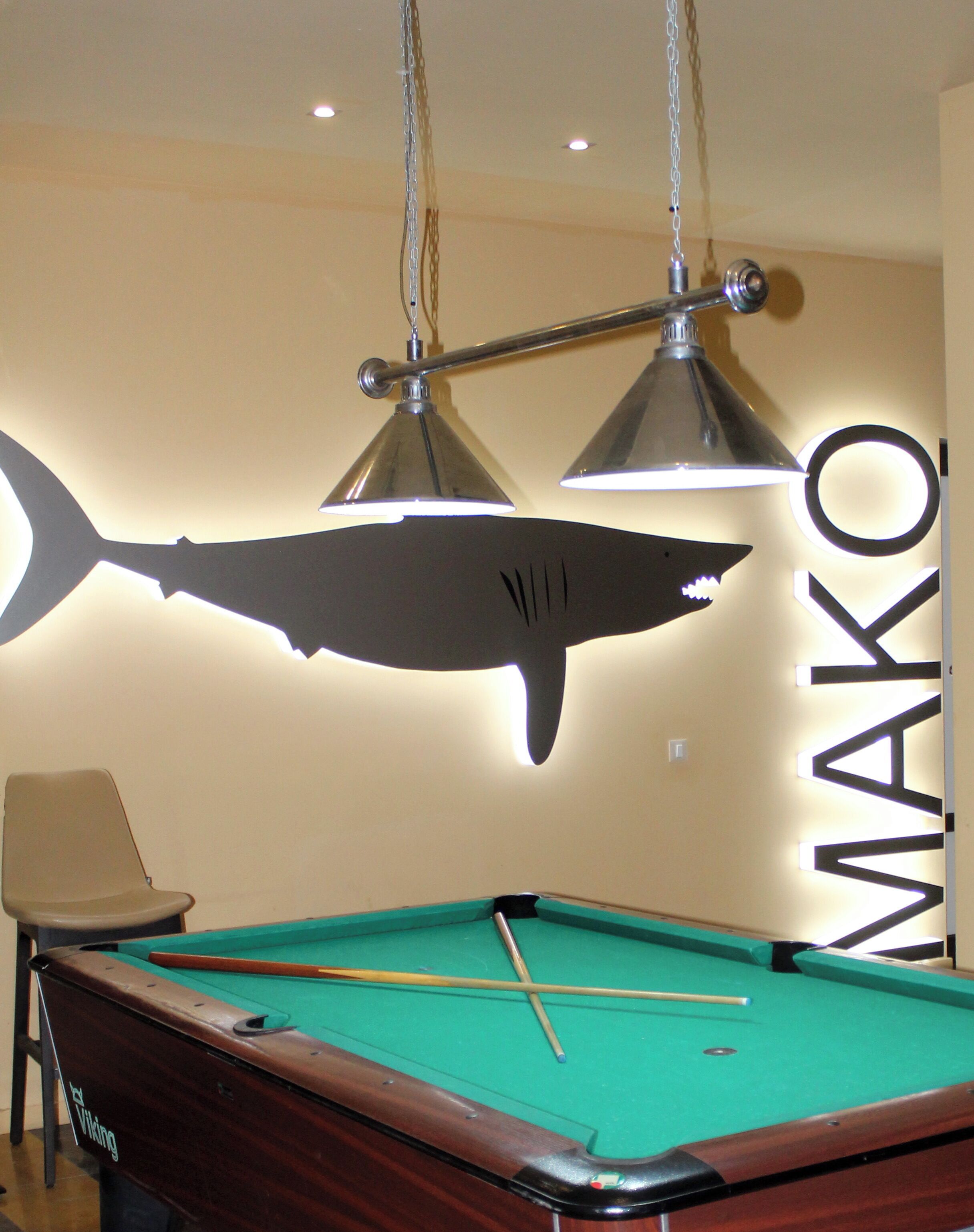 A pool table with a shark decoration, a chair, and overhead lighting fixtures.