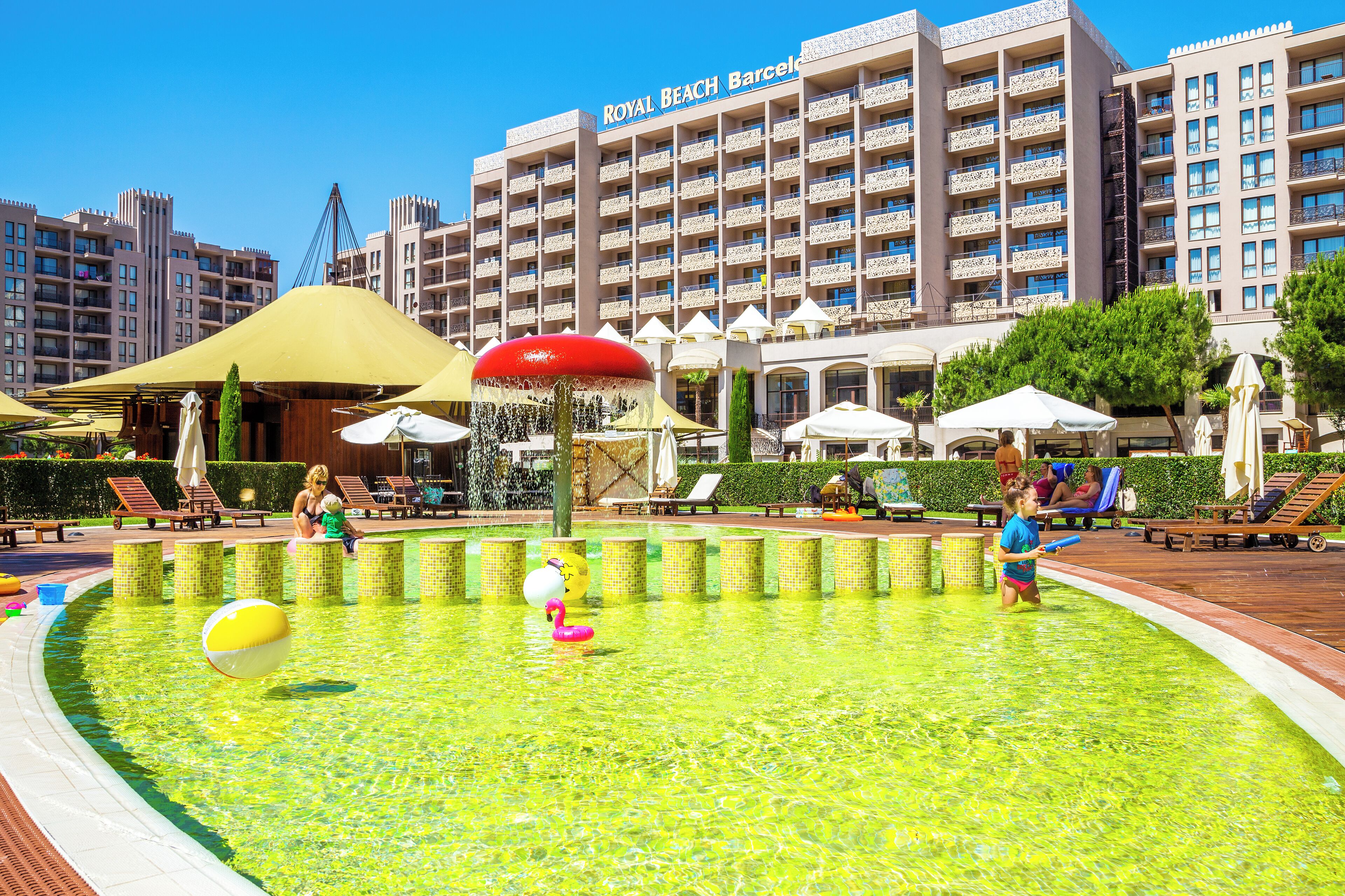 Zona de piscina amb gandules i ombrelles, una font d'aigua i un edifici al fons amb la façana que diu "Royal Beach Barcelona".