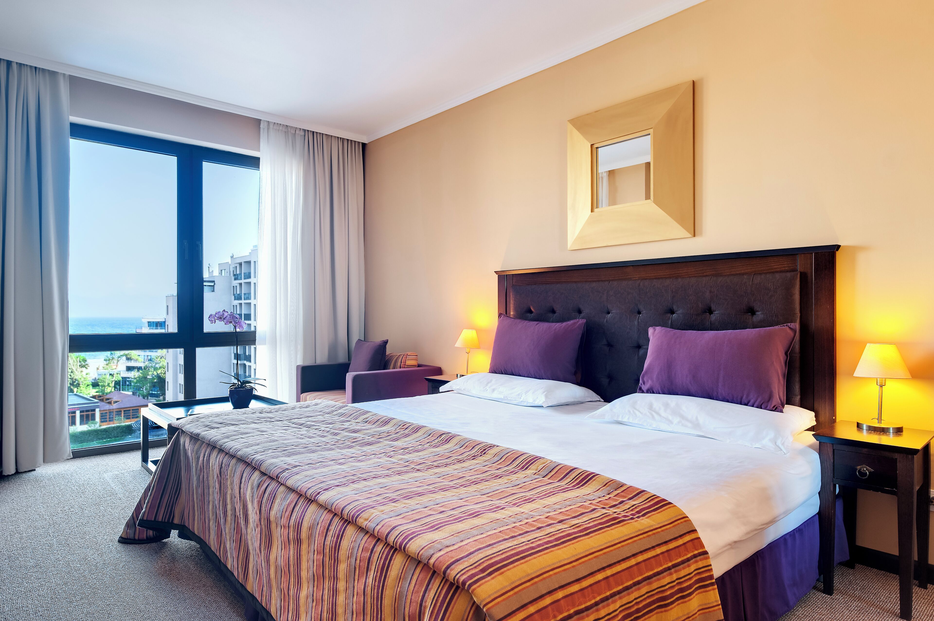 A hotel room with a large bed, a nightstand, a chair, a window with a view of buildings, and a potted plant.