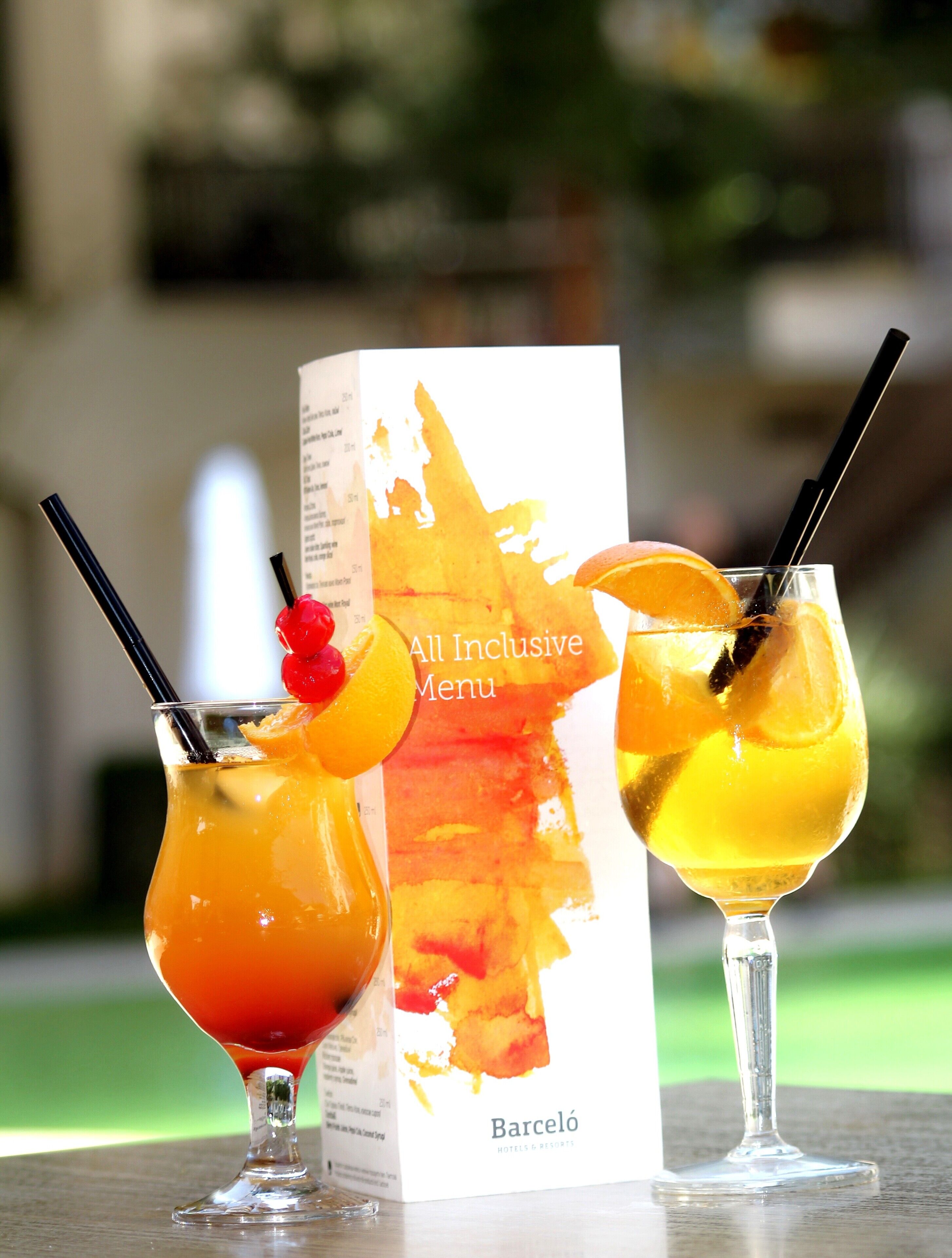 Two cocktails with garnishes and straws, a menu, and the word "Barceló" on a table.