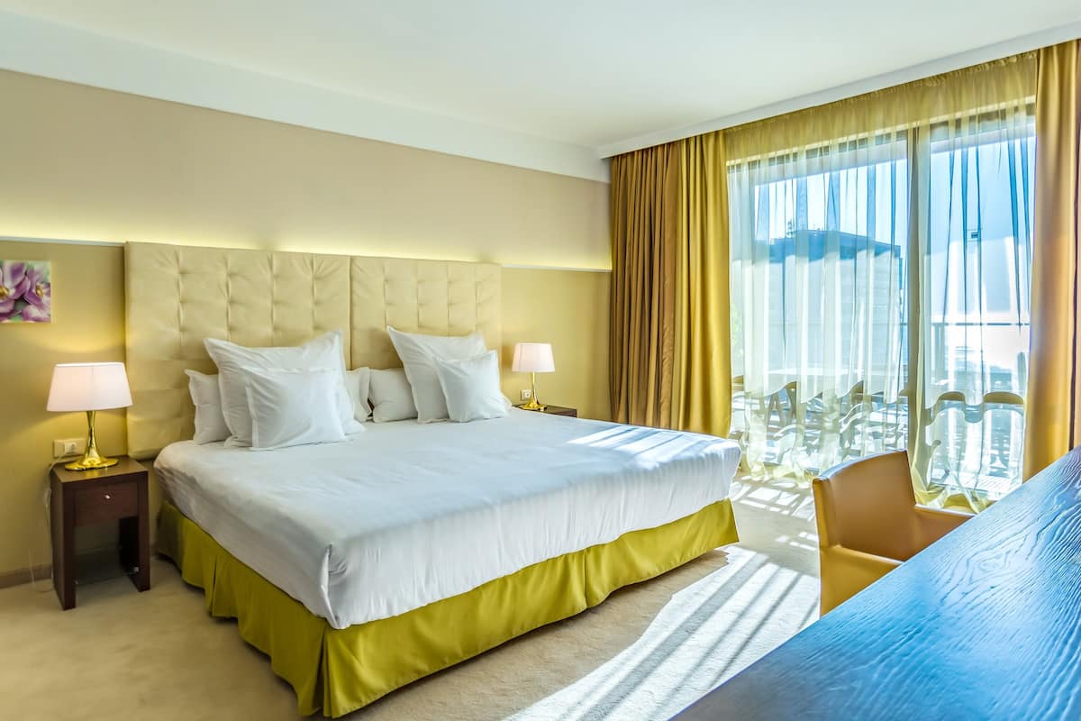 A hotel room with a large bed, two bedside tables with lamps, a view of the city through large windows, and a chair by the window.
