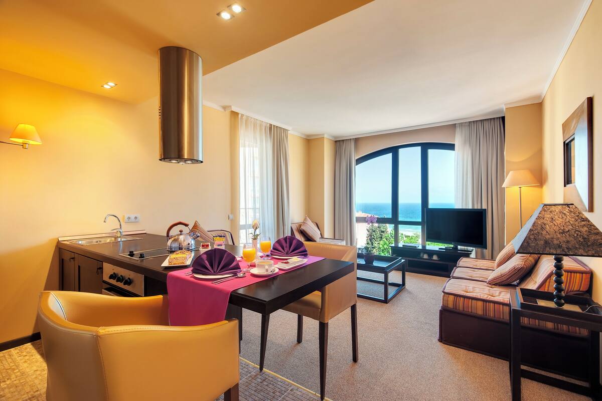 A hotel room with a dining area, a sofa, a TV, and a balcony with a view.