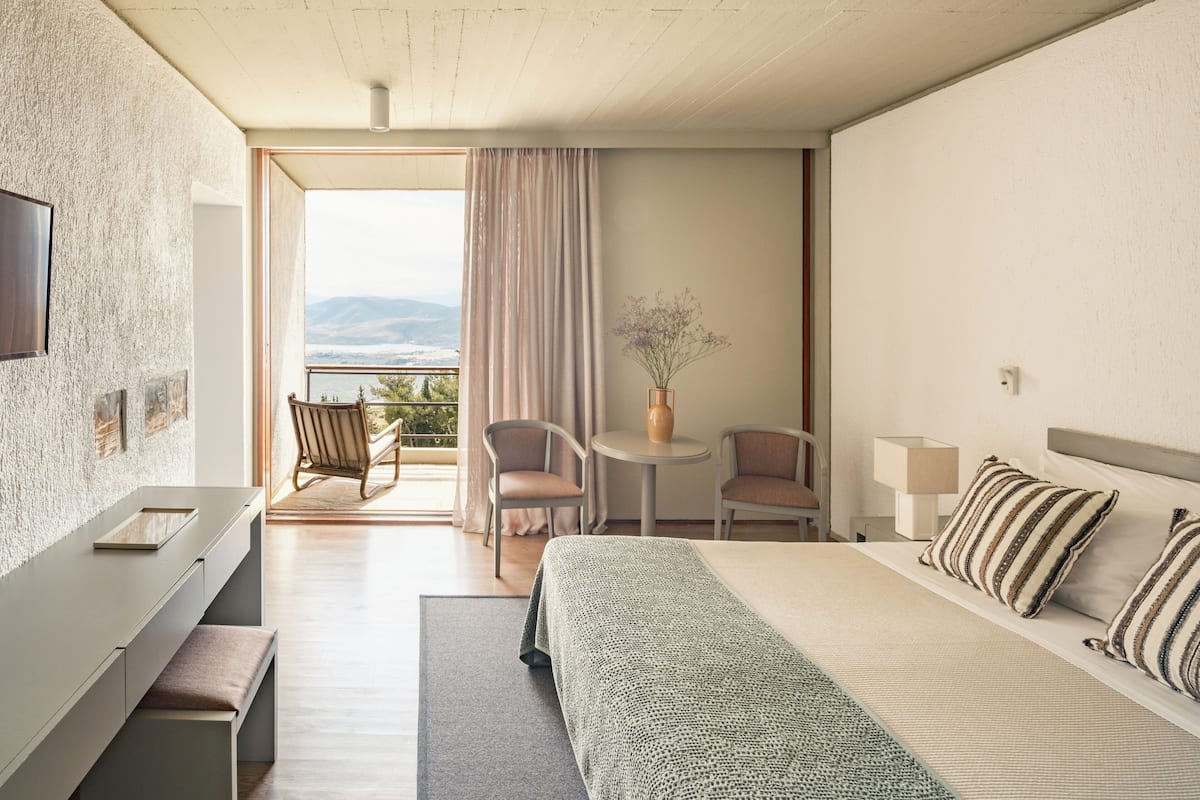 A hotel room with a bed, a desk, a chair, a small table with a vase, and a view of the outdoors.