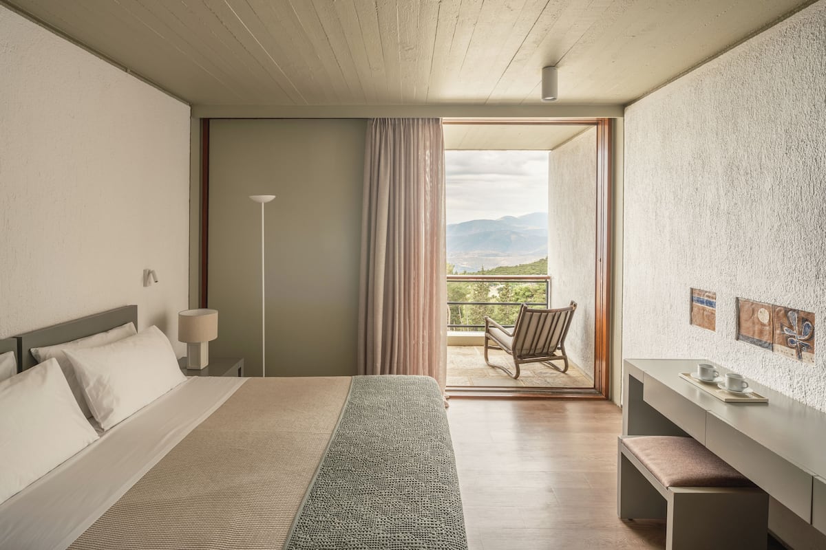 A hotel room with a bed, a desk, a chair, and a balcony with a view of the mountains.