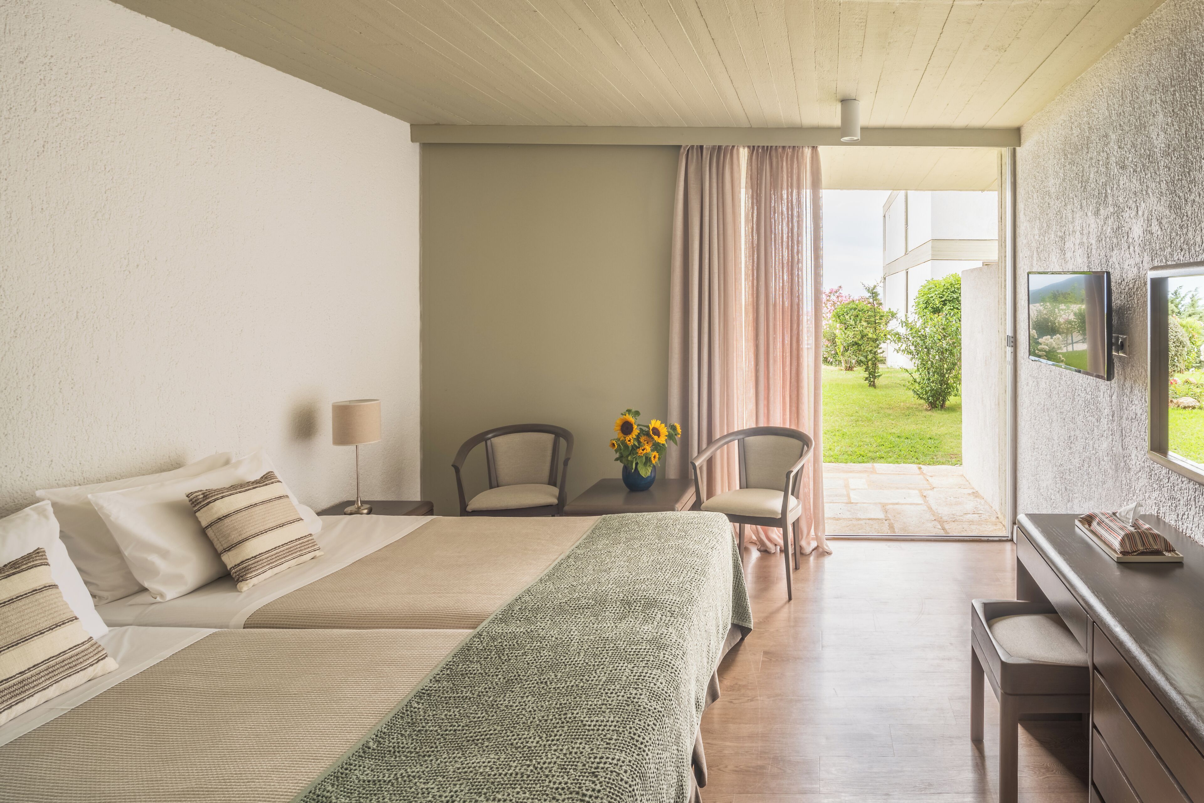 A hotel room with two beds, a desk, chairs, a TV, and a view of the garden.