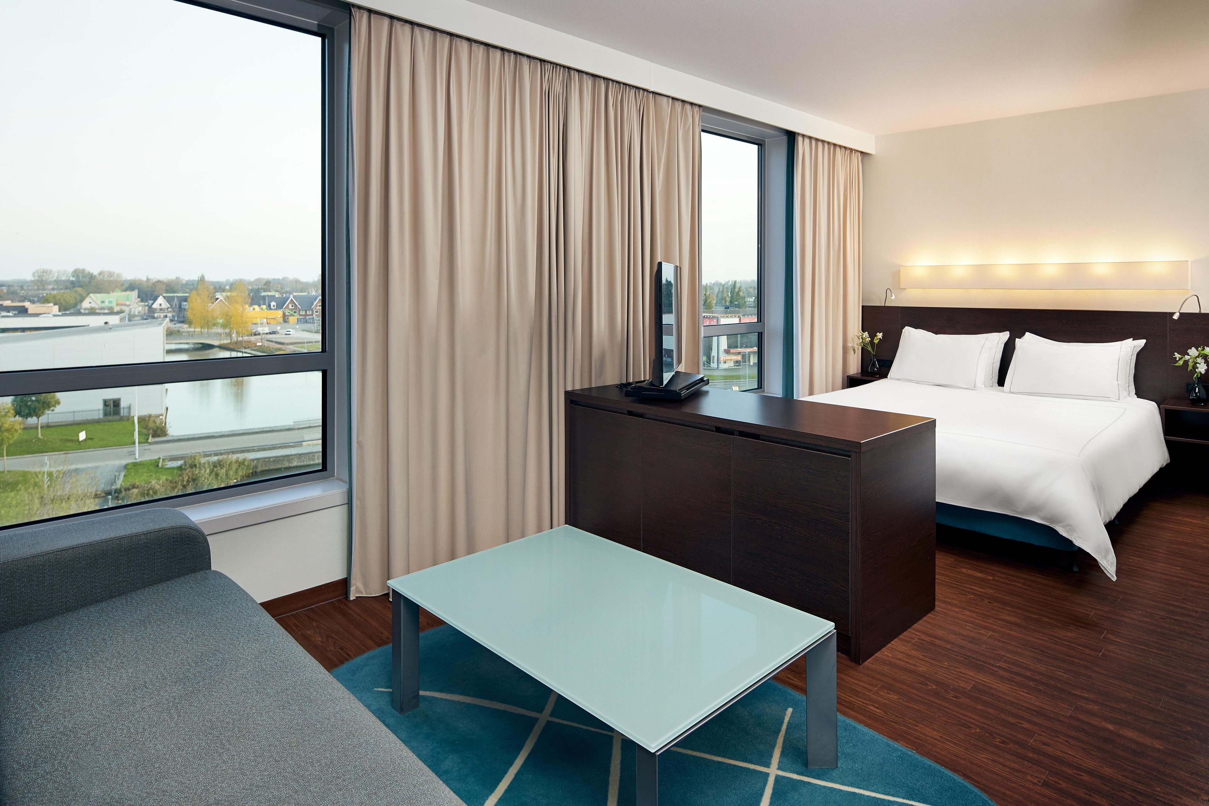 A hotel room with a bed, a desk, a chair, a TV, and a view of a cityscape.