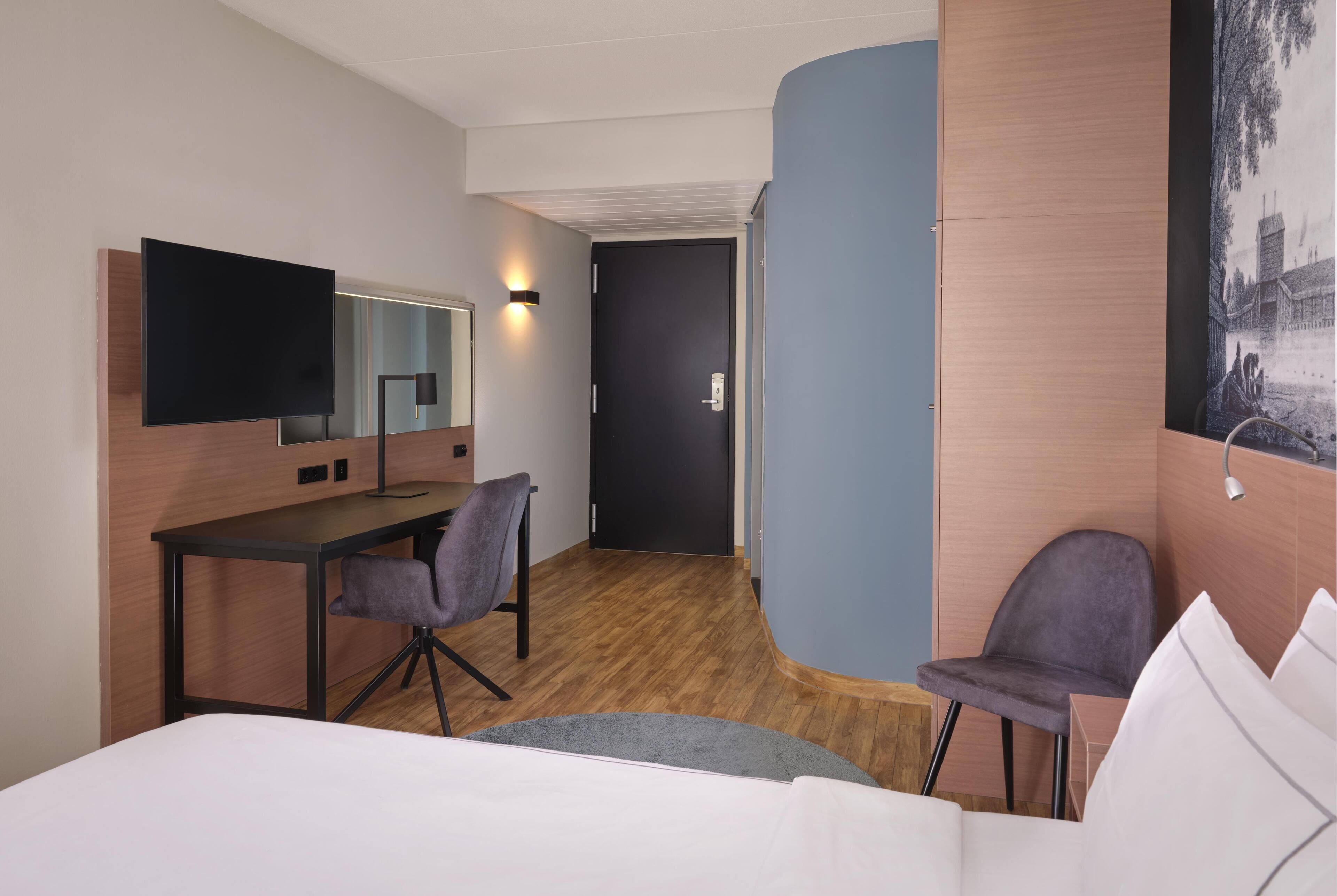 Una camera d'albergo con un letto, una scrivania, una sedia, una TV, una porta e un quadro appeso al muro.