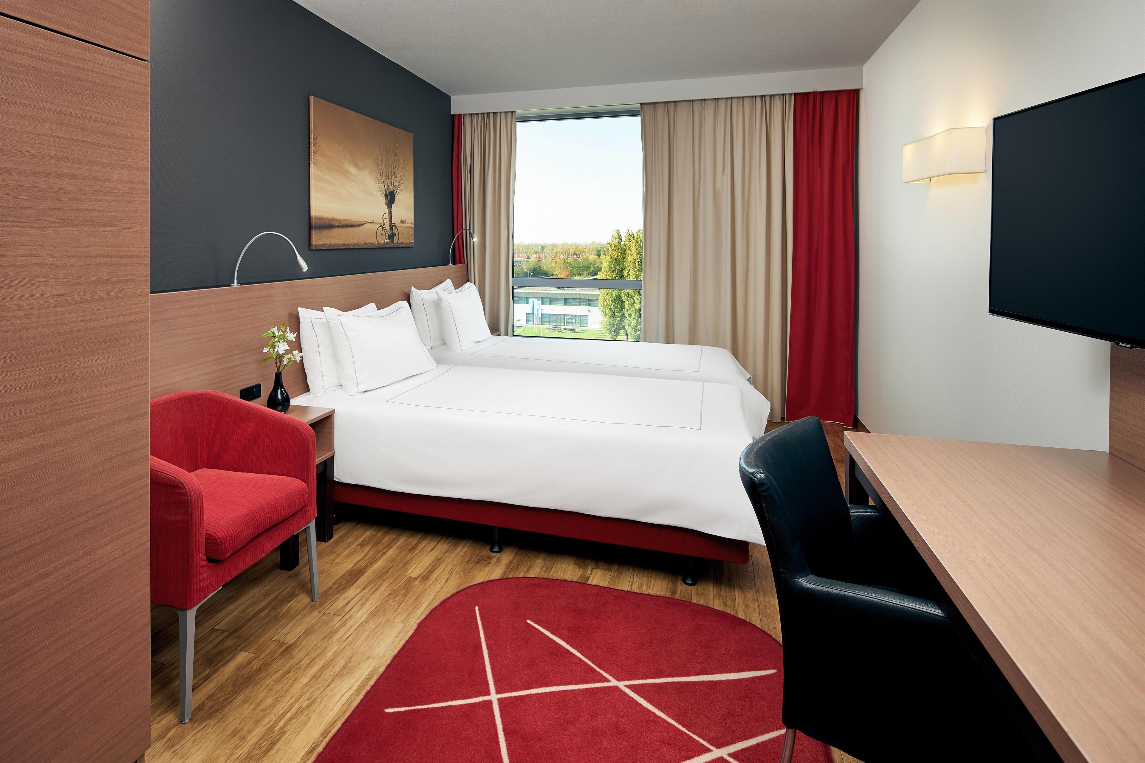 Une chambre d’hôtel avec un lit, un bureau, une chaise, une télévision, un tableau et une fenêtre avec des rideaux.