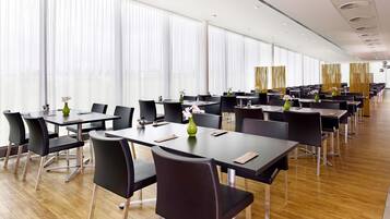 Een vergaderzaal met meerdere tafels en stoelen, grote ramen met dunne gordijnen en een modern plafond.