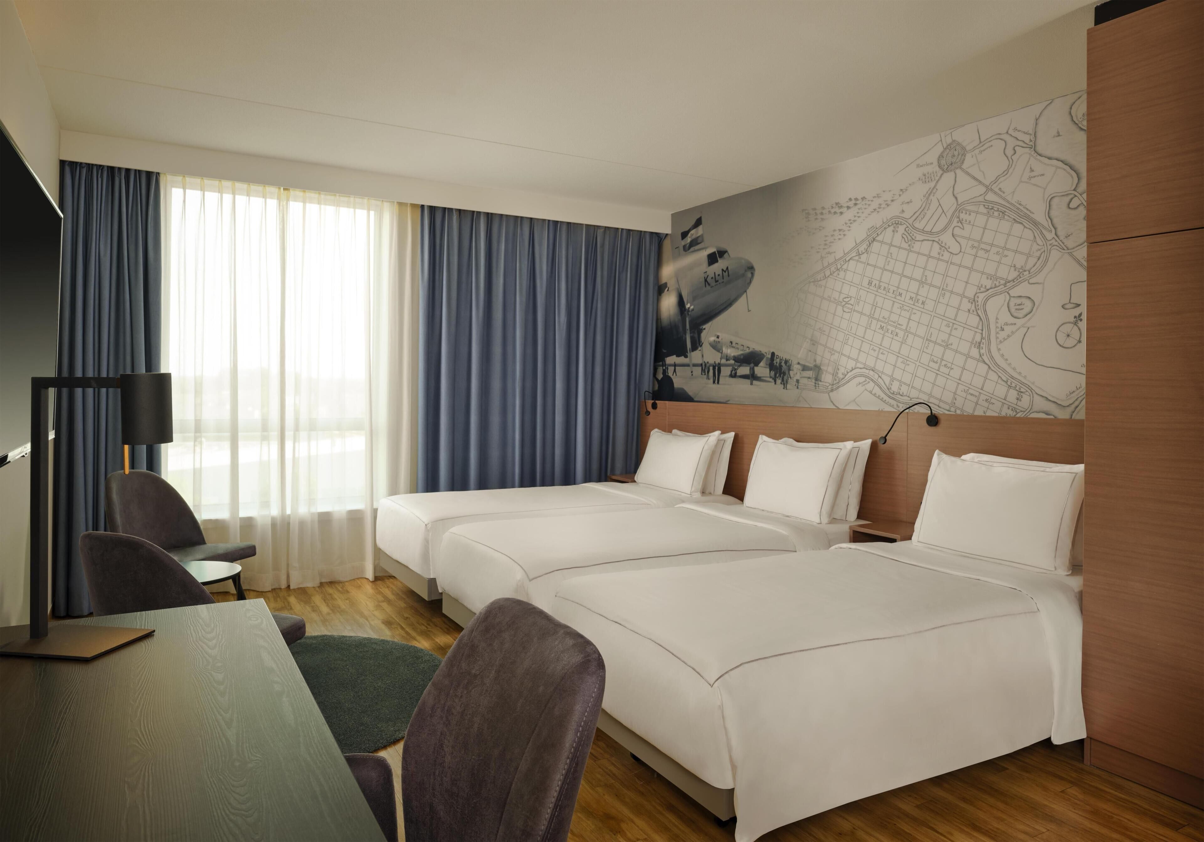 A hotel room with two beds, a desk, a chair, a map on the wall, and a window with curtains.