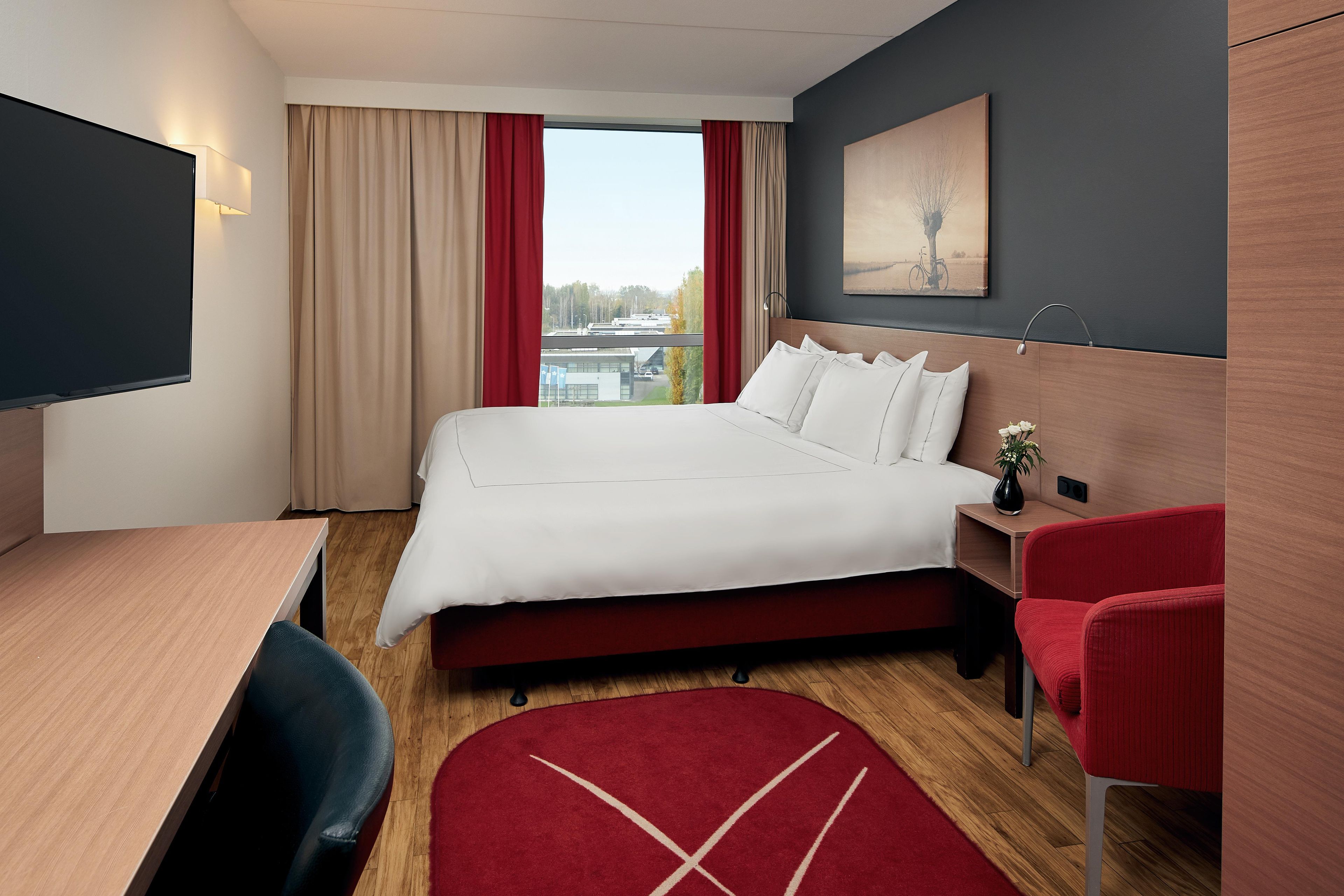Una habitación de hotel con cama, escritorio, silla, televisor y ventana con cortinas.