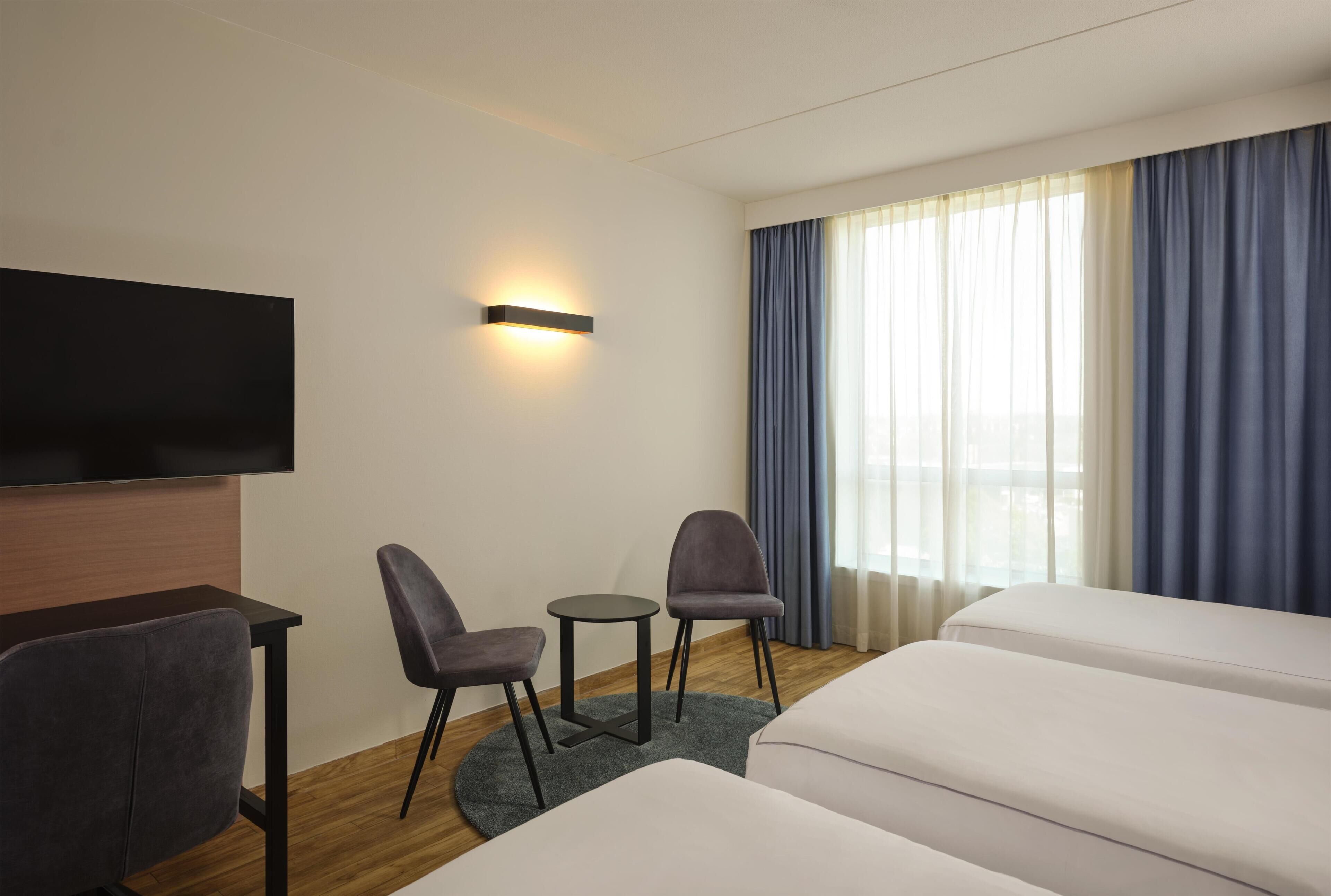 Ein Hotelzimmer mit einem an der Wand befestigten Flachbildfernseher, zwei Sesseln, einem kleinen runden Tisch, einer Couch und einem großen Fenster mit Vorhängen.