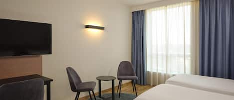 Ein Hotelzimmer mit einem an der Wand befestigten Flachbildfernseher, zwei Sesseln, einem kleinen runden Tisch, einer Couch und einem großen Fenster mit Vorhängen.
