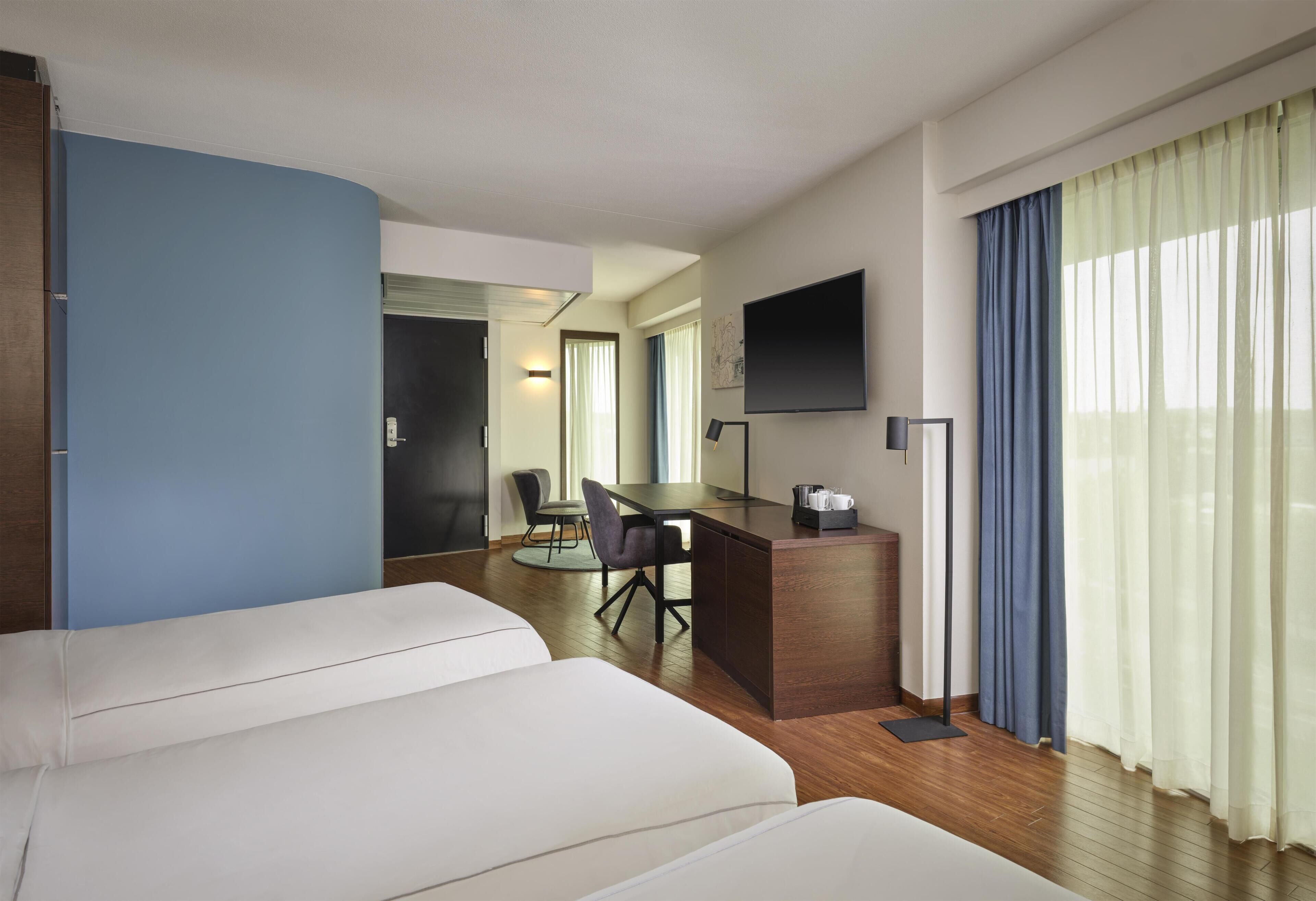 A hotel room with a bed, a desk with a chair, a flat-screen TV, and a window with curtains.