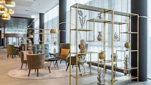 A modern interior with a gold-framed shelving unit displaying decorative items, plush seating, and a large window providing natural light. - Park Plaza Amsterdam Airport (Lijnden)