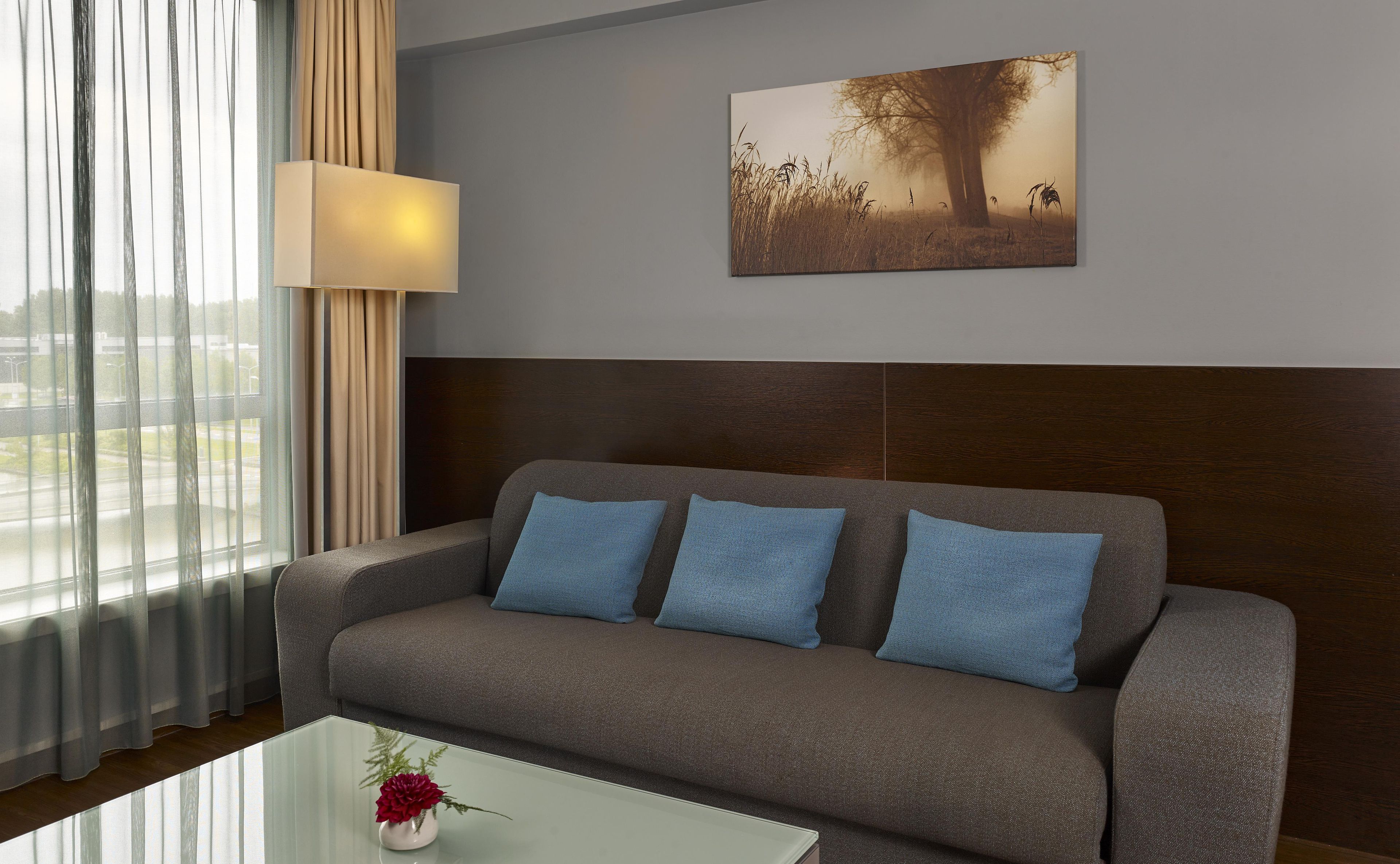 A modern living room with a grey sofa, blue cushions, a glass coffee table with a flower vase, a painting of a tree and grass, and sheer curtains.