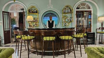 Zona de bar con un cantinero, mostrador de madera y sillas tapizadas de verde.