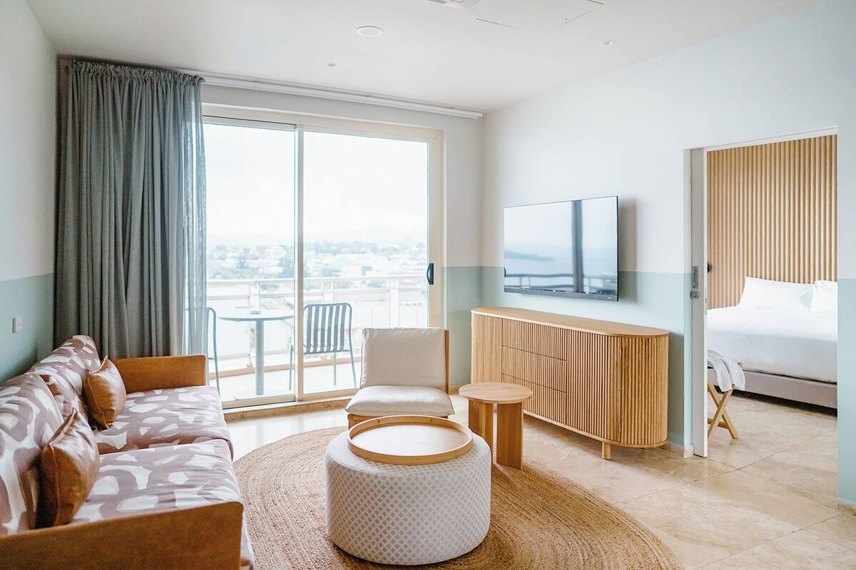 A modern hotel room with a sofa, ottoman, TV, and a bed.