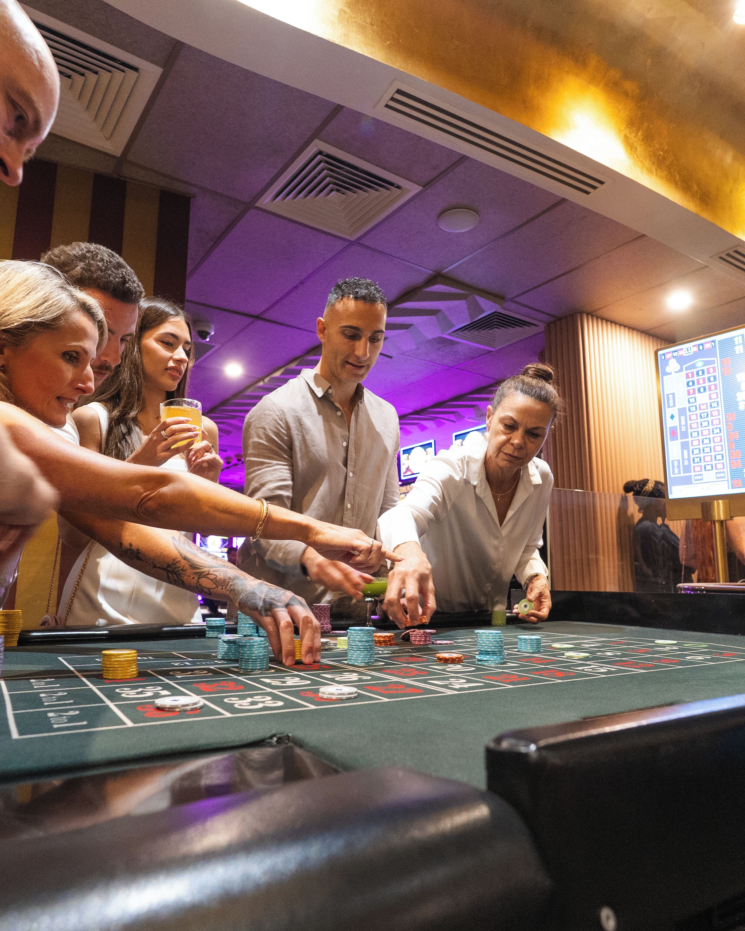 A group of people playing blackjack at a casino table with a roulette wheel visible in the background.