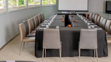 Un salón de conferencias con una larga mesa, sillas, una pantalla de proyector y un amplio ventanal con vistas a zonas verdes.