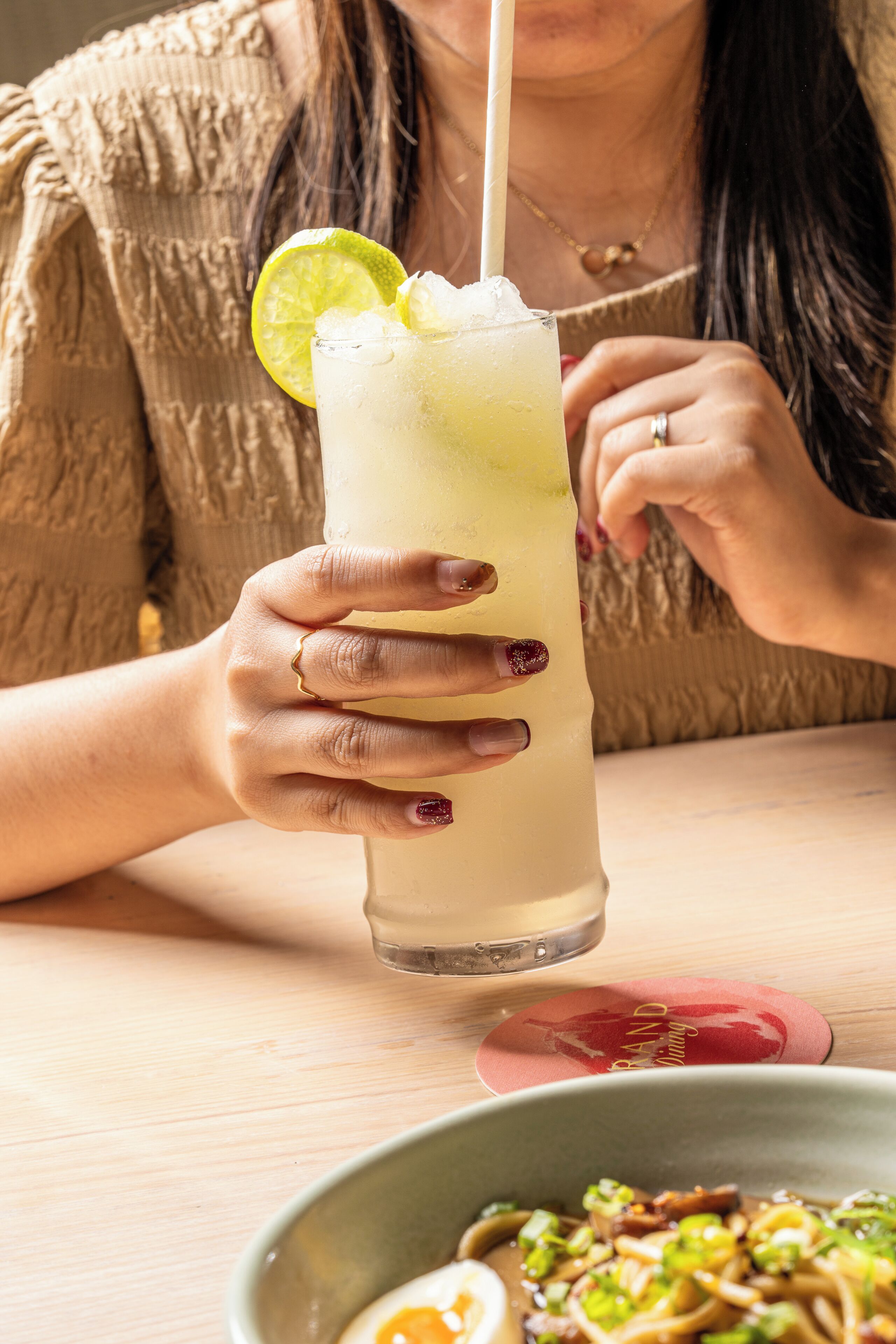 A person holding a tall glass with a straw, a lime wedge, and ice cubes.