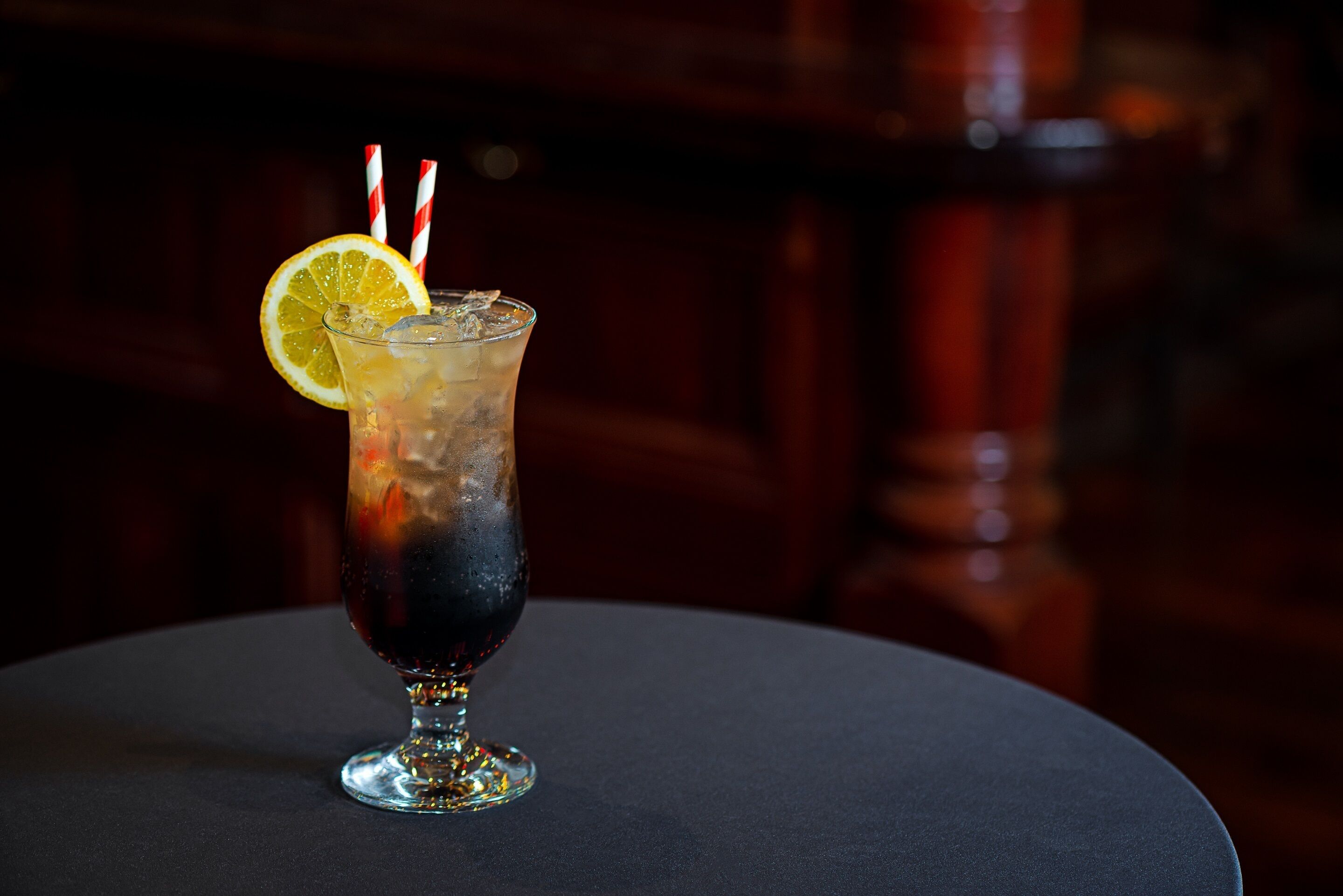 A cocktail glass with a lemon slice and striped straws on a round table.