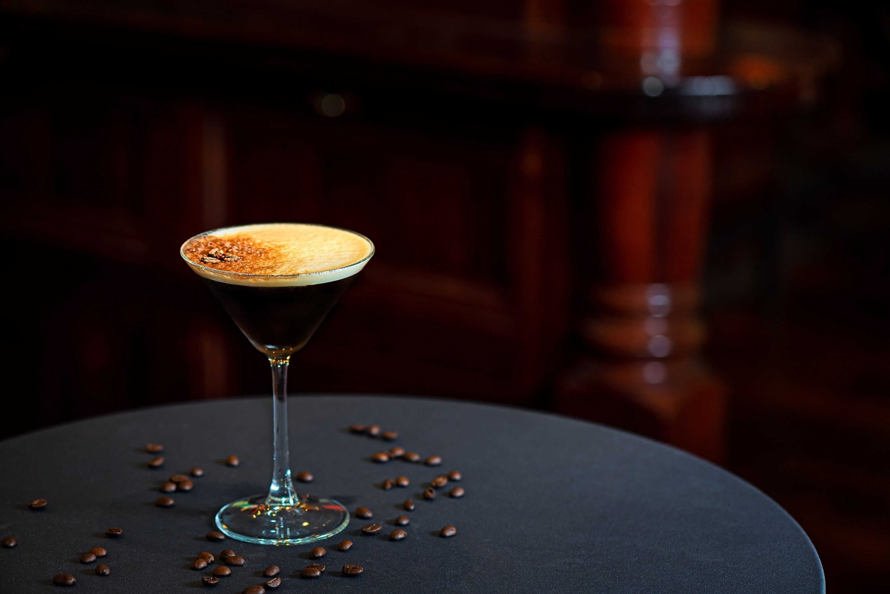 A martini glass with a frothy top on a round table surrounded by coffee beans.