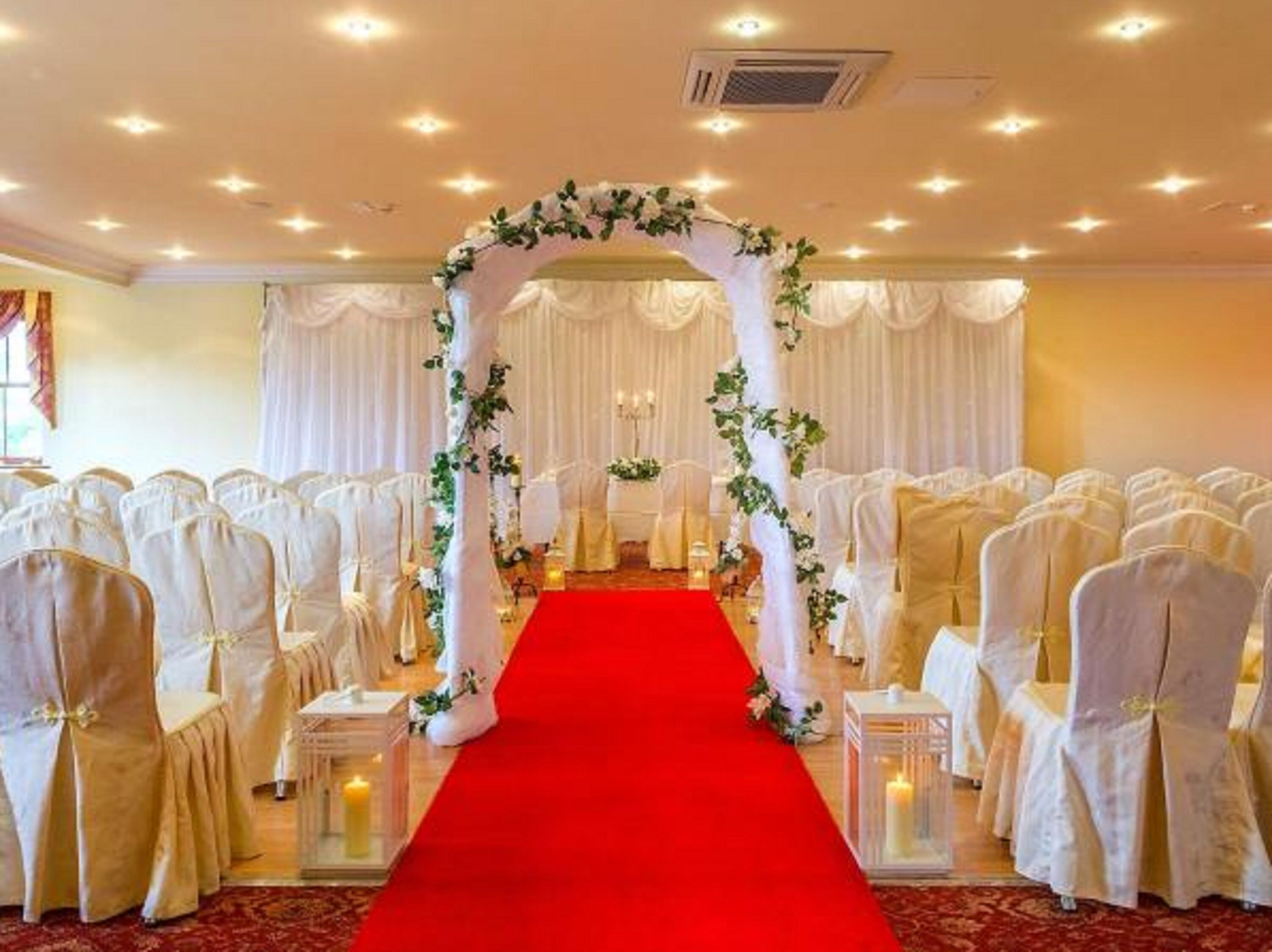 Un pasillo de bodas con sillas blancas y una alfombra roja, que conduce a un arco decorado.