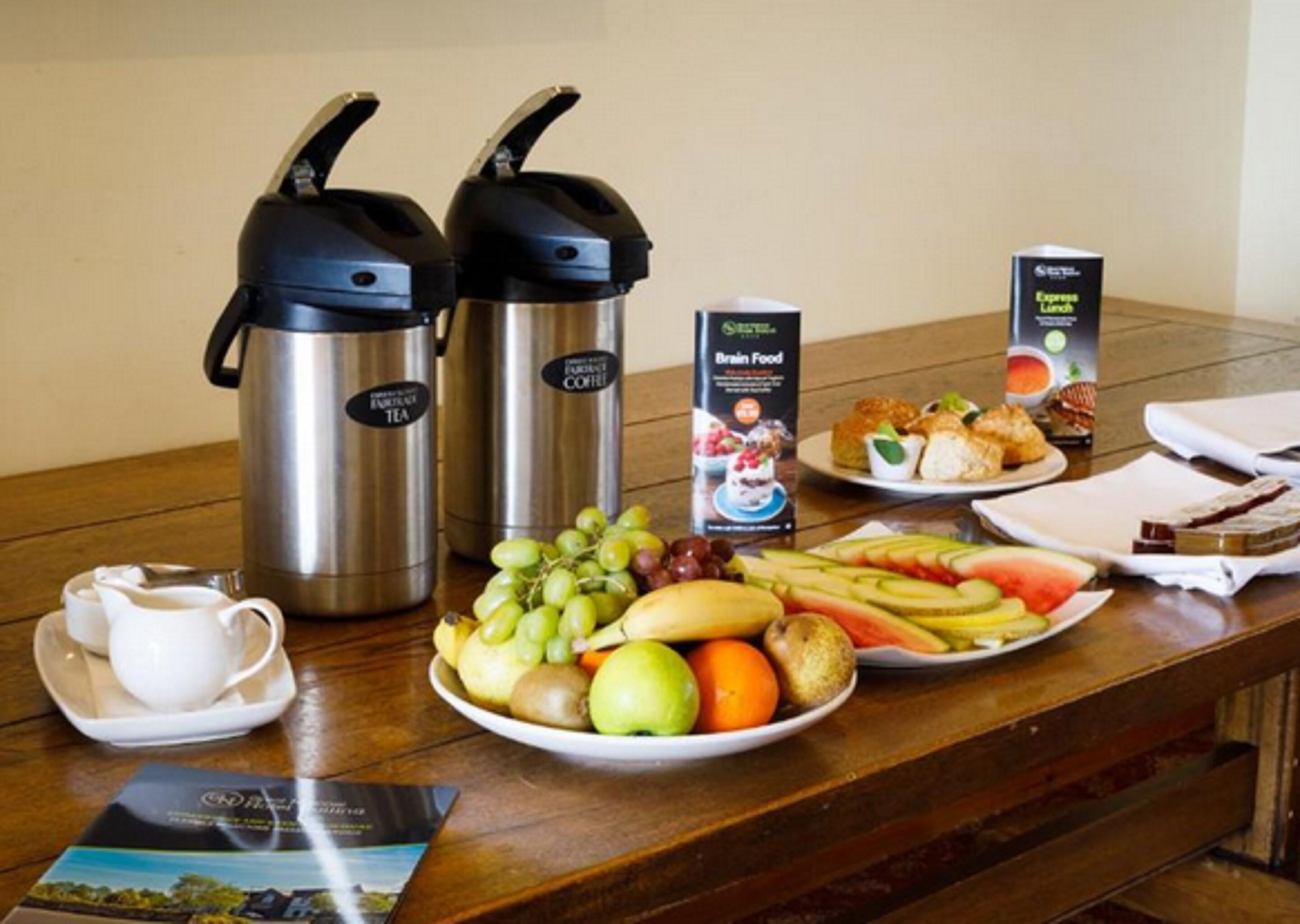 A breakfast setup with two stainless steel thermos cups, a carton of brain food, a plate of fresh fruits, a cup of coffee, and a brochure on the table.