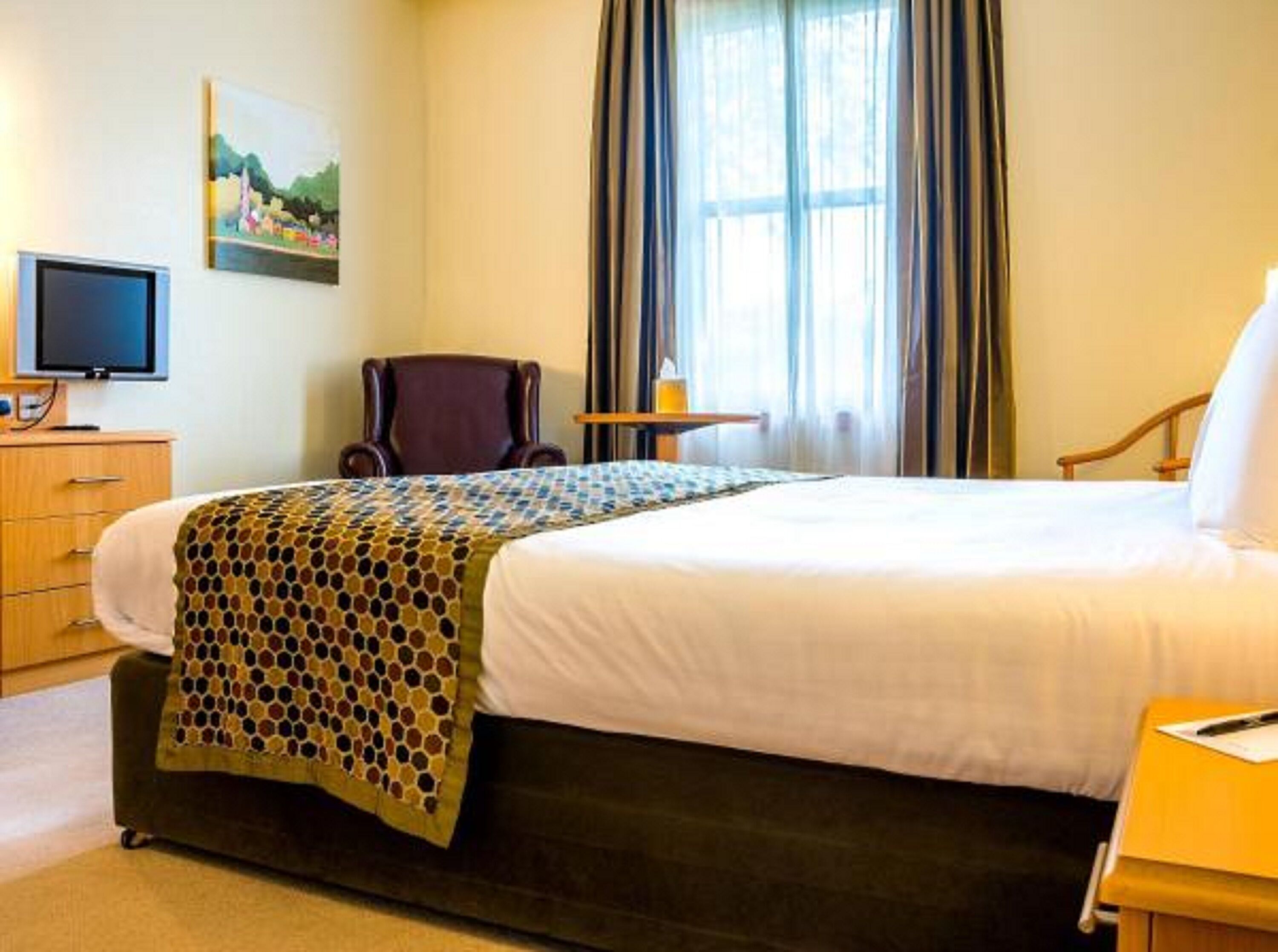 A hotel room with a bed, a chair, a desk, a television, and a painting on the wall.
