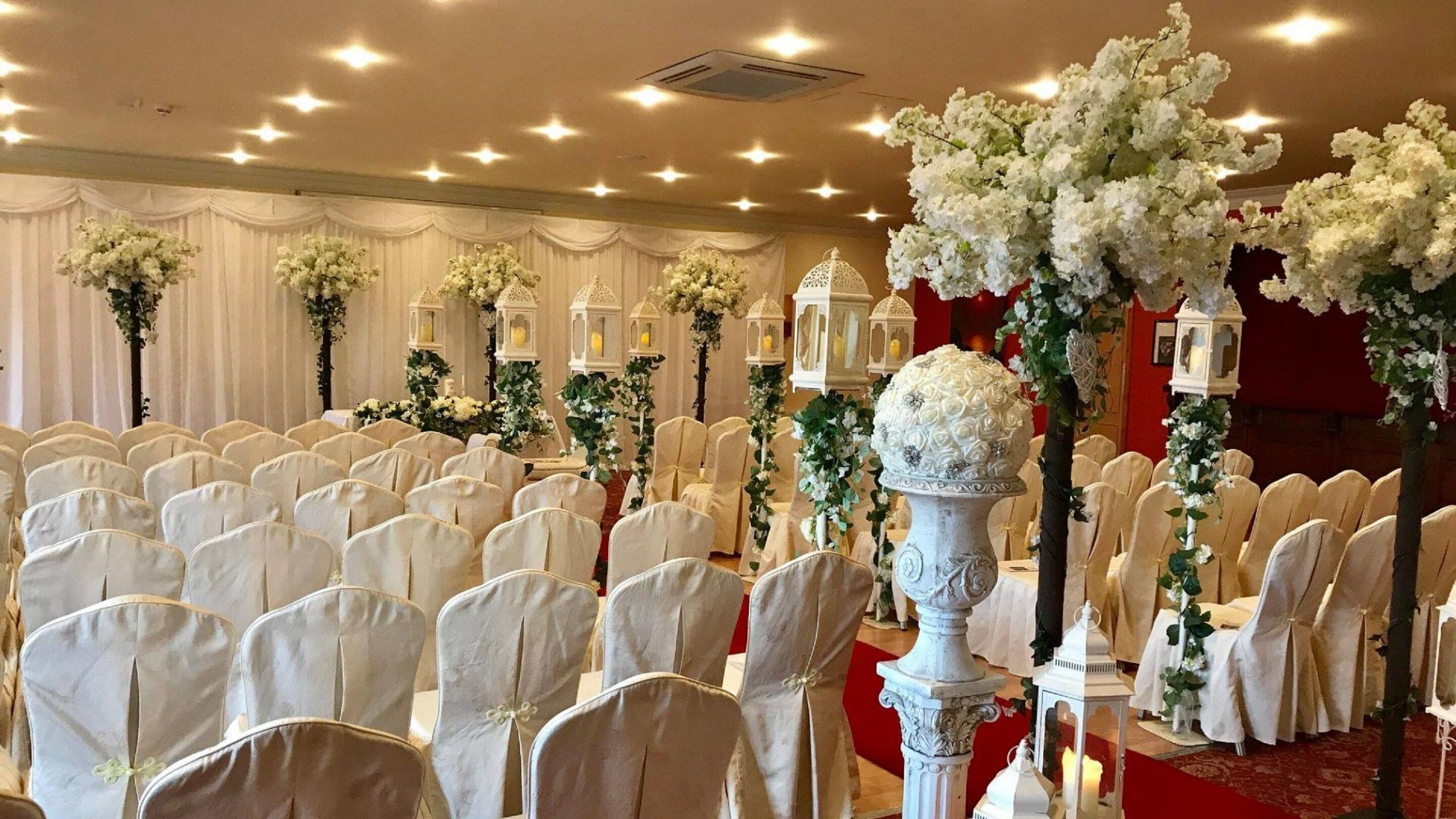 Un salón de banquetes con filas de sillas blancas, arreglos florales blancos y faroles blancos.
