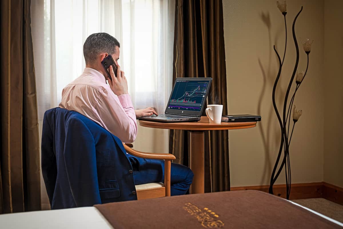 Een man in een roze overhemd die een smartphone en een laptop gebruikt, met een aandelenmarkt grafiek op het scherm, zittend aan een klein, rond tafeltje met een witte mok.