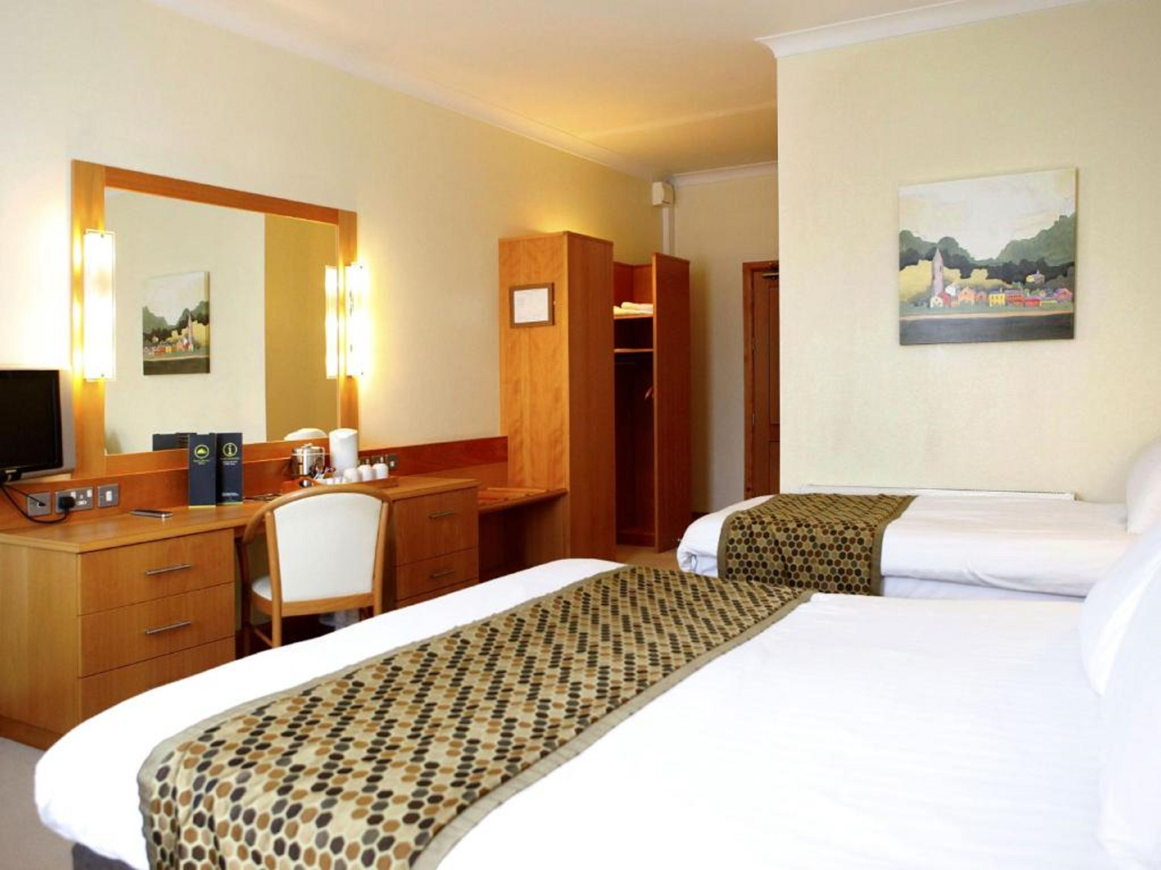 Ett hotellrum med två sängar, ett skrivbord med en stol, en tv, en spegel och en tavla på väggen.