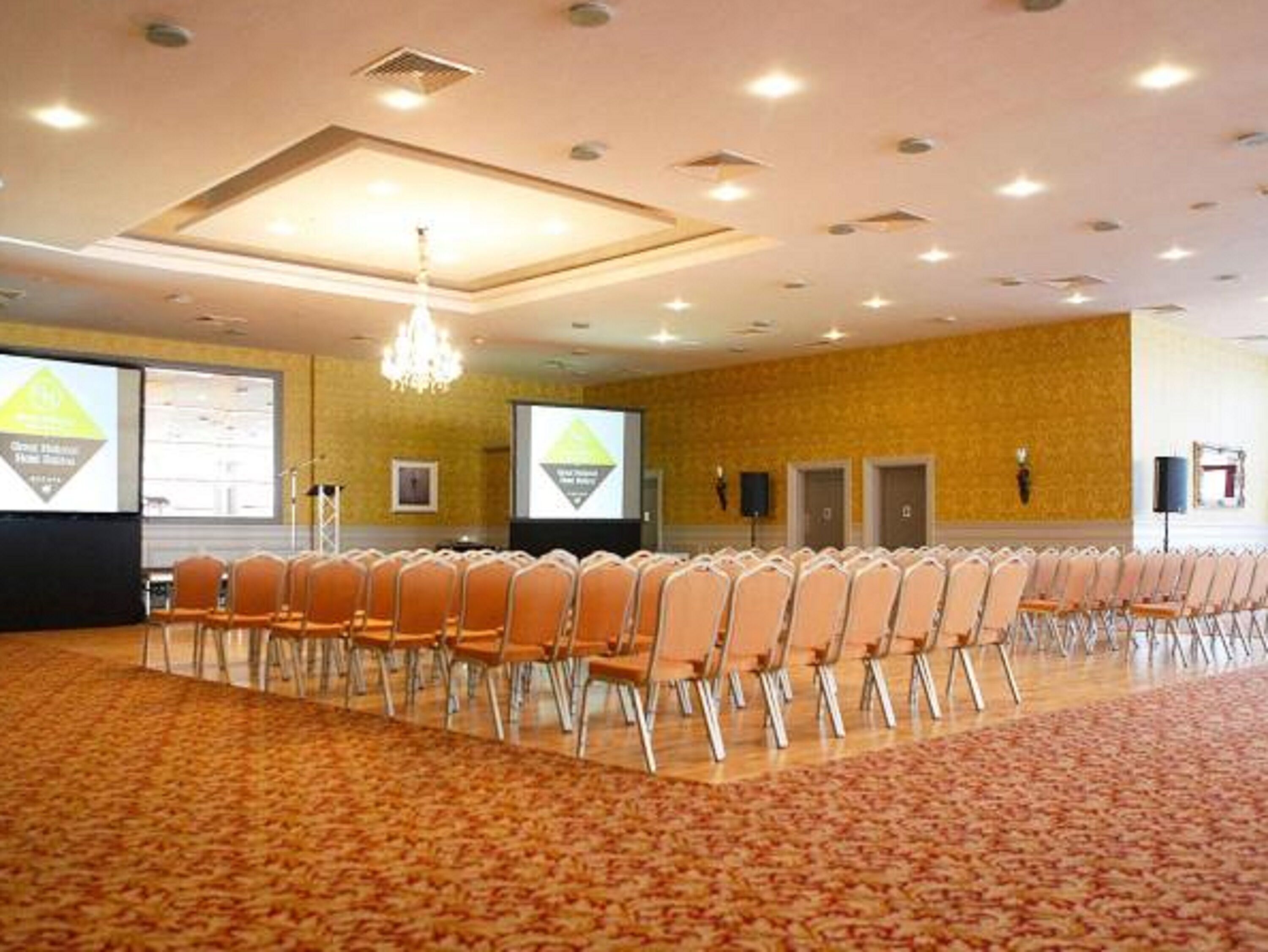 A conference room with rows of chairs, a carpeted floor, a chandelier, and a projector screen.